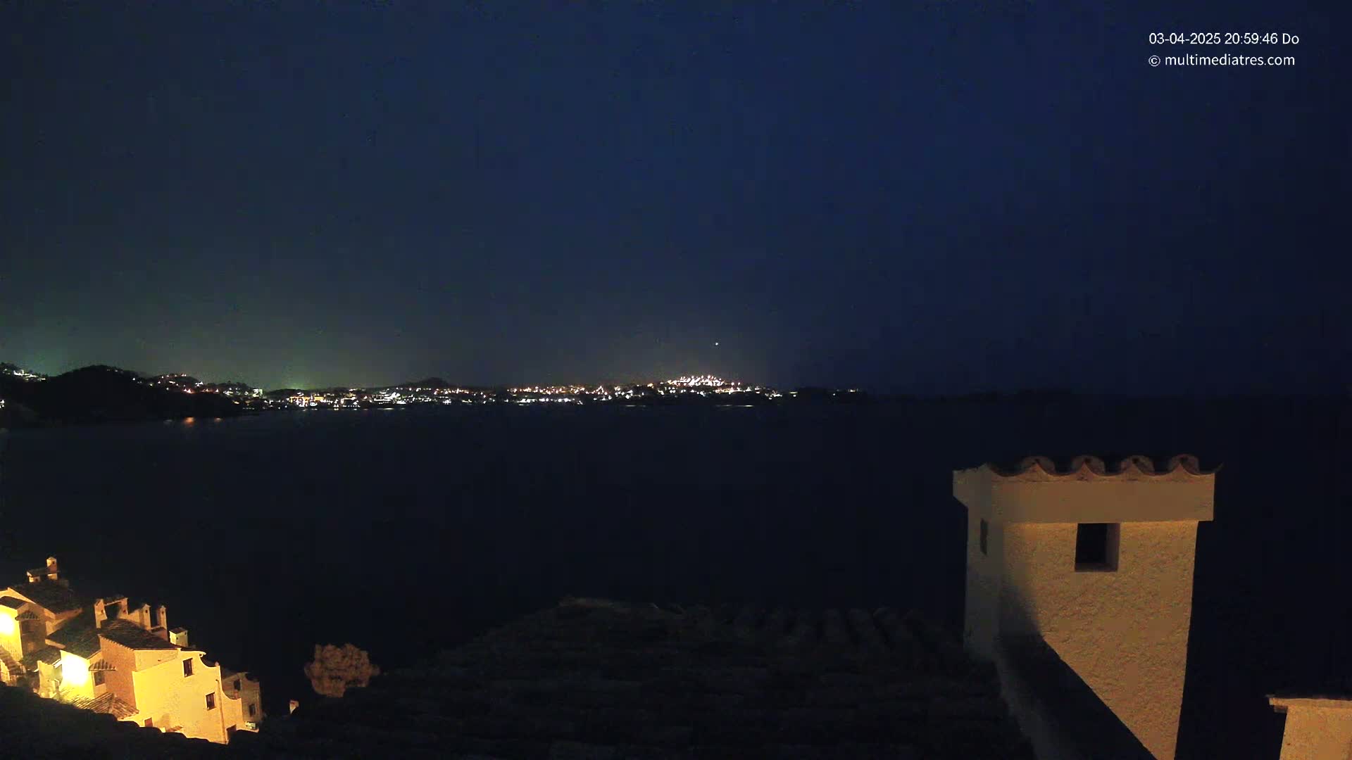 A nighttime view of a town's lights across a dark body of water, seen from a hilltop with buildings illuminated in the foreground.
