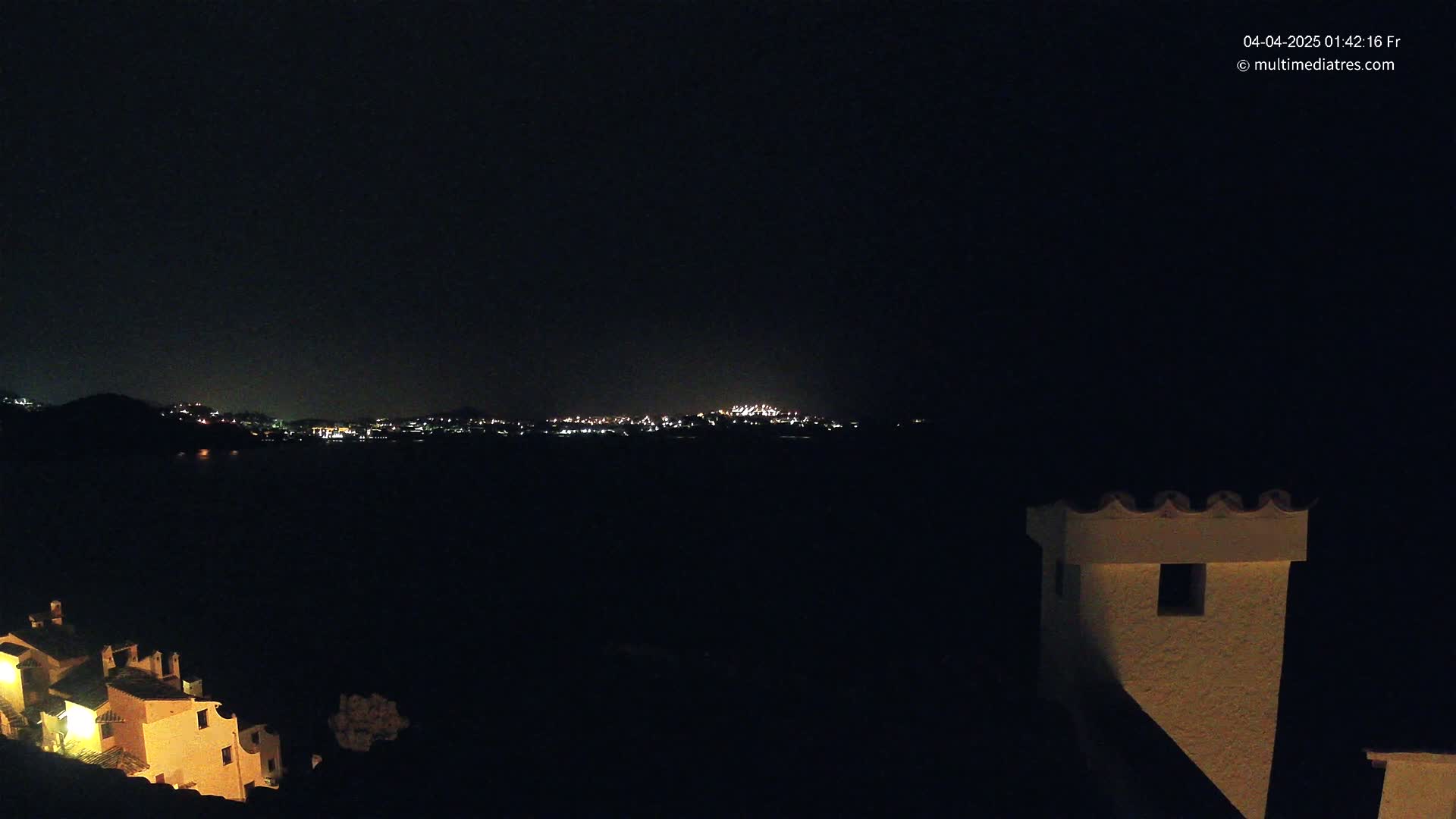 A nighttime view of a town's lights across a dark body of water, partially obscured by a hill, with illuminated buildings in the foreground.