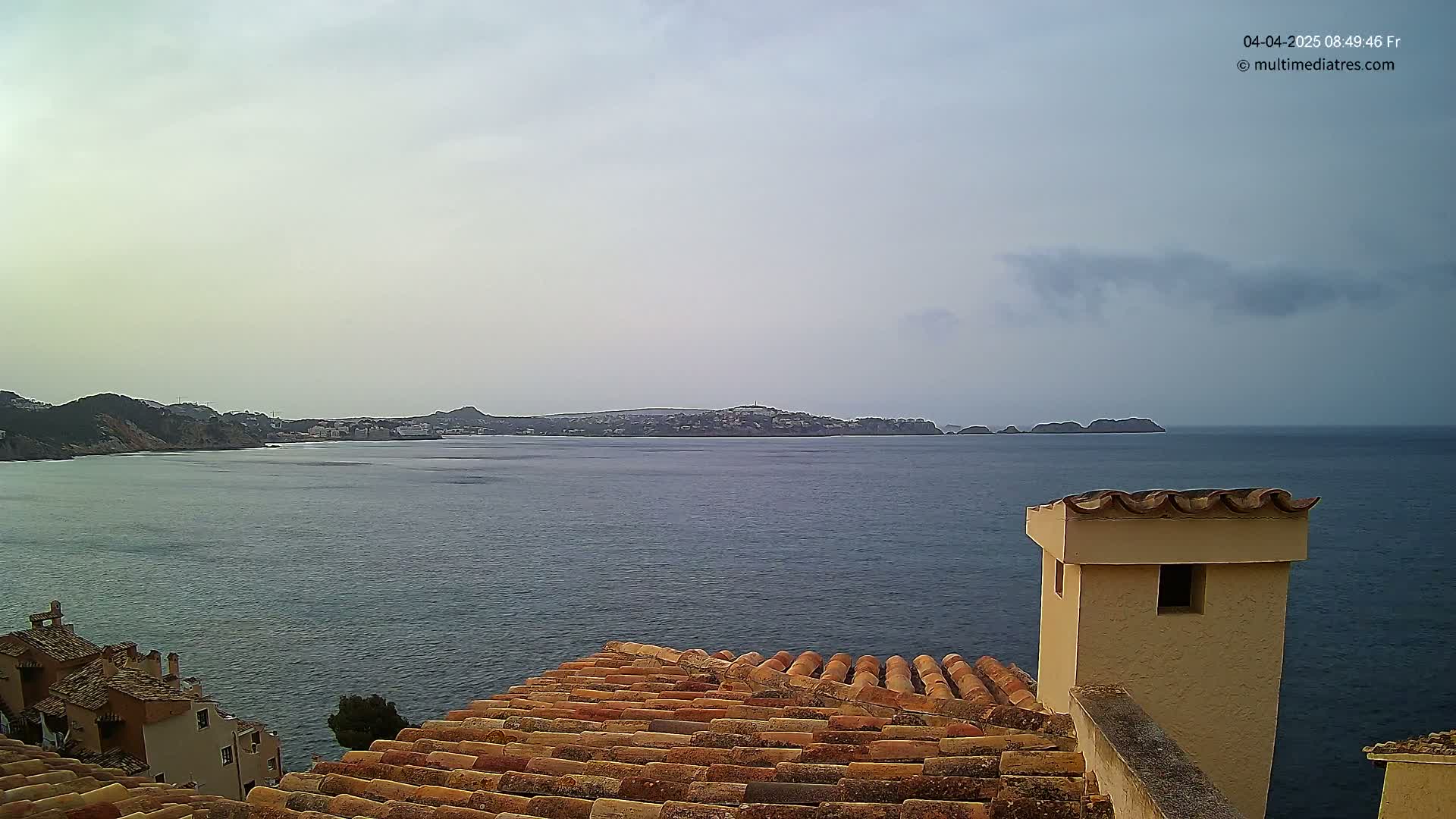 A view from a rooftop with terracotta tiles overlooking a calm sea and distant hills under a mostly cloudy sky.