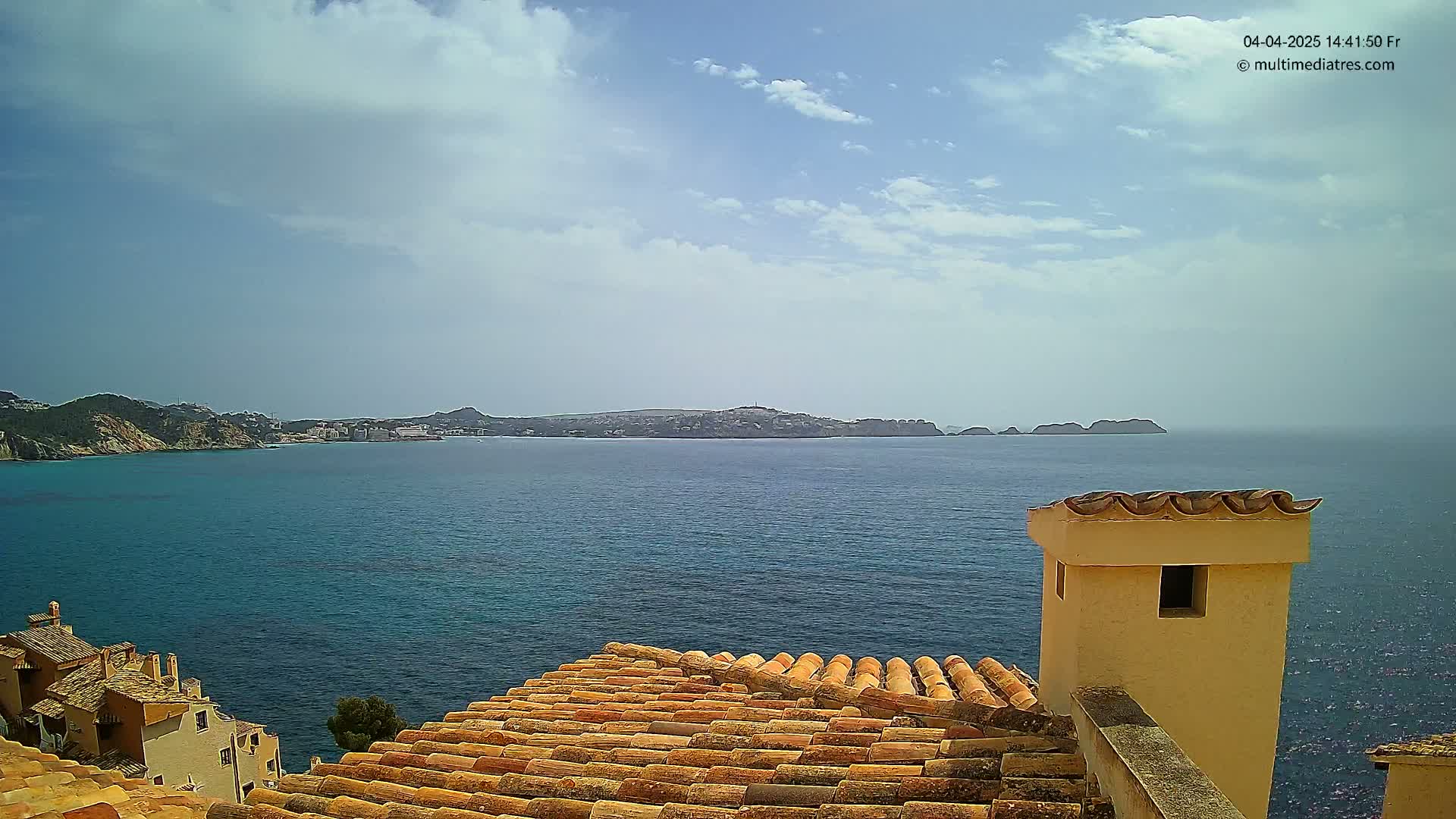 A partly cloudy day overlooking a calm blue sea, with a distant island and a portion of a terracotta-tiled roof in the foreground.