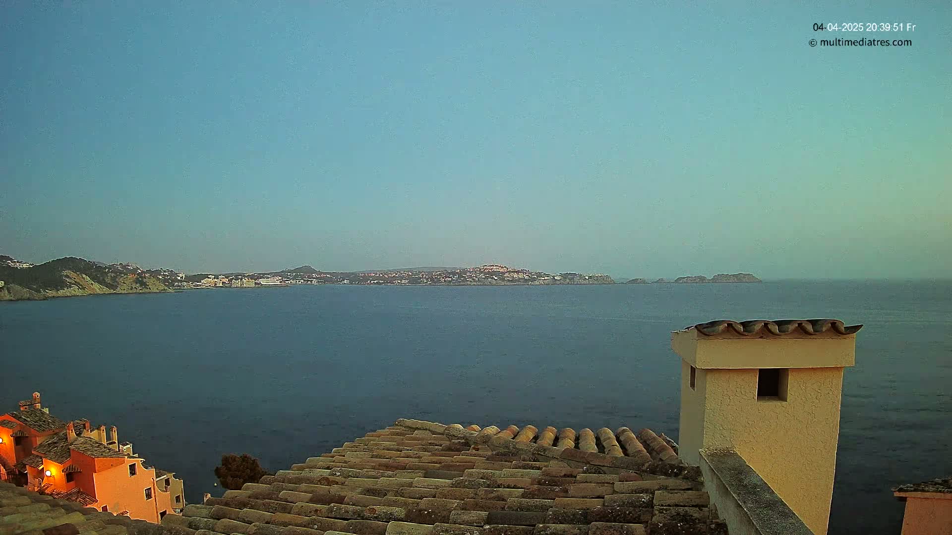 A calm, dark-blue sea stretches to a distant coastline under a clear twilight sky, viewed from a tiled roof with a light-beige chimney.