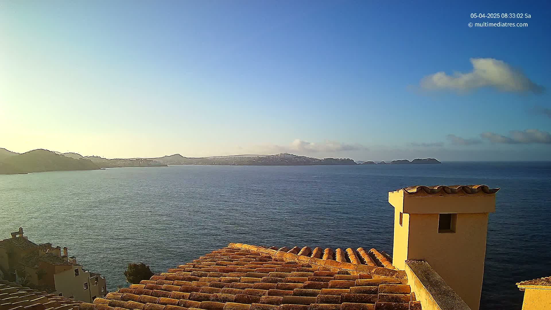 A calm, blue ocean stretches to a distant, hilly coastline under a mostly clear sky with a few small clouds, viewed from a terracotta roof with a light beige chimney.