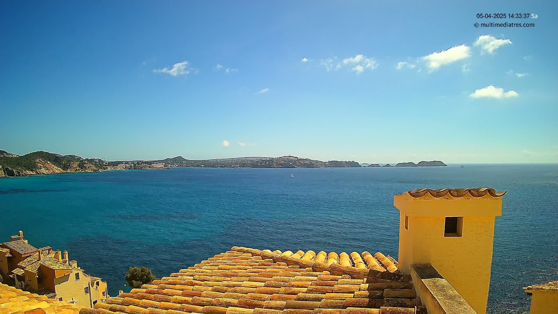 A coastal view on a sunny day shows a terracotta roof overlooking a calm blue sea and distant hills.