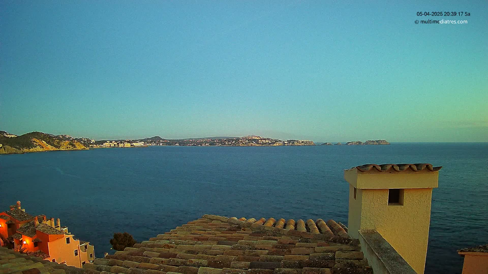 A calm, clear blue sea stretches to a distant coastline under a pale blue sky, viewed from a tiled roof with a light-colored chimney.