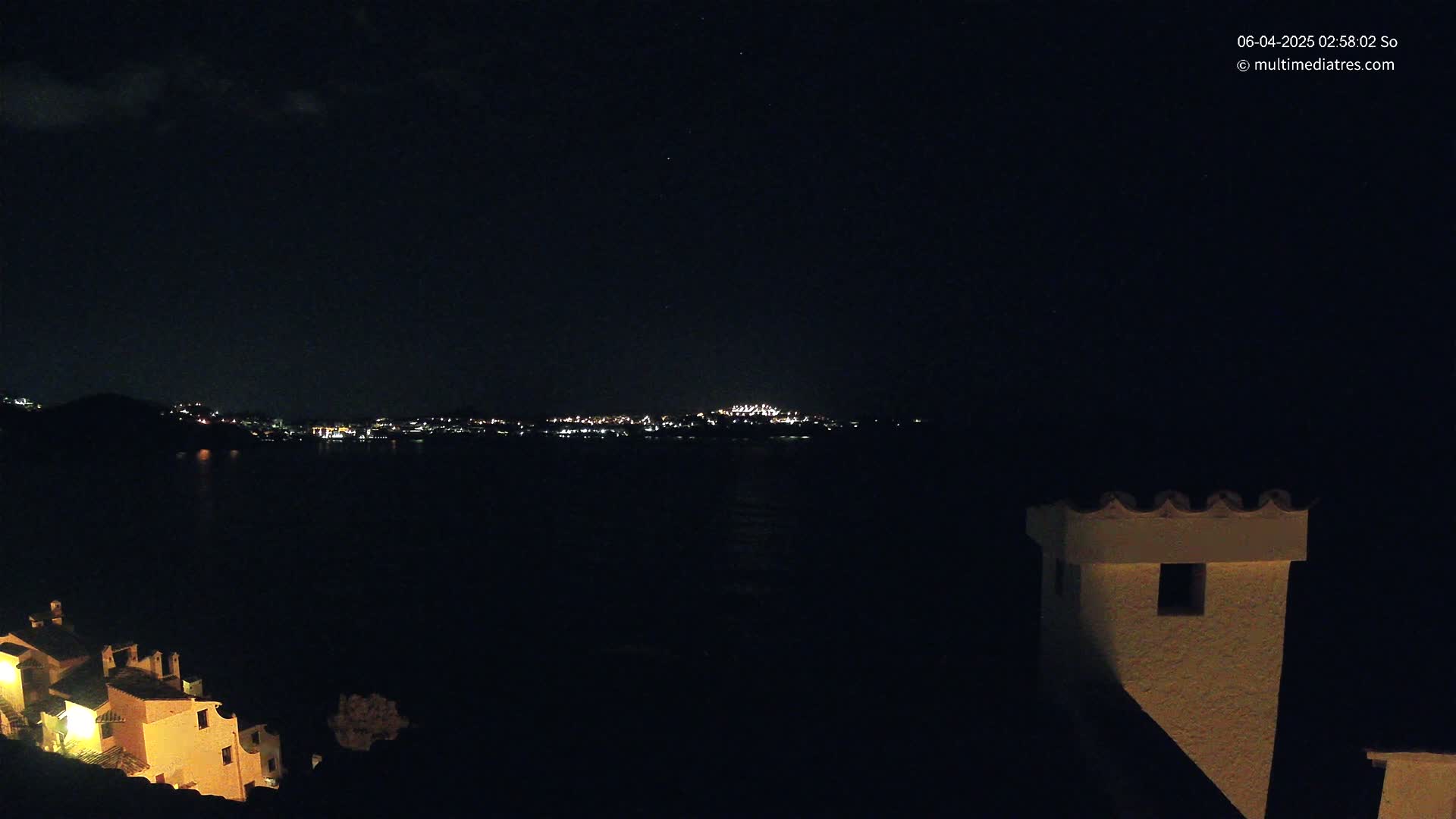 A nighttime view shows a dark body of water separating a dimly lit town from a hillside with illuminated buildings, under a mostly clear night sky.