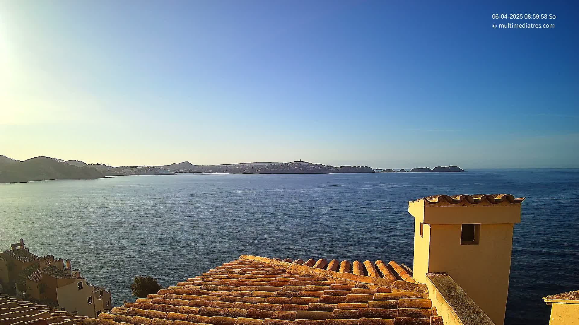 A terracotta roof overlooks a calm, blue sea with distant hills and islands under a clear blue sky.