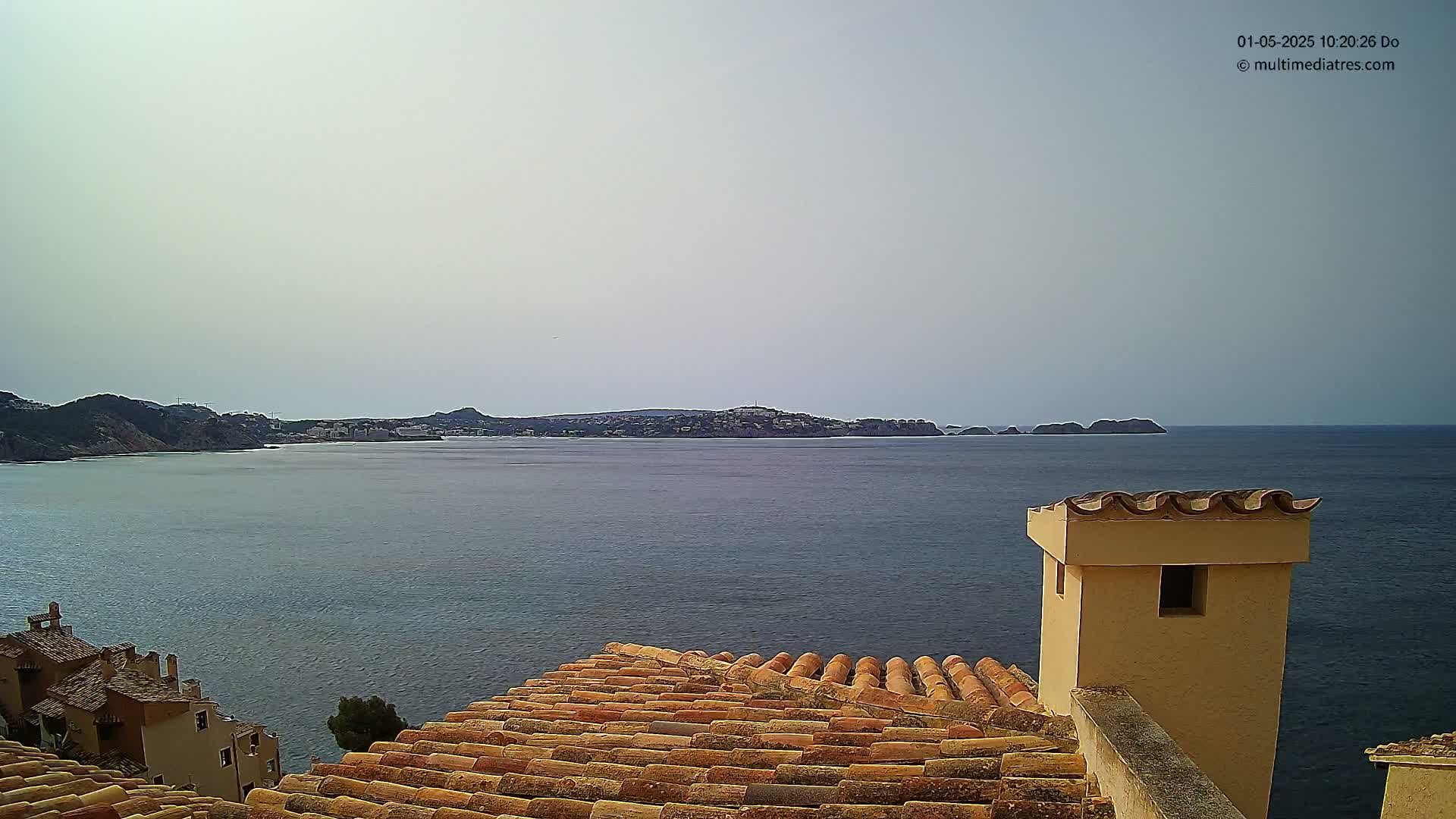 A tiled rooftop overlooks a calm sea and distant coastline under a hazy sky.