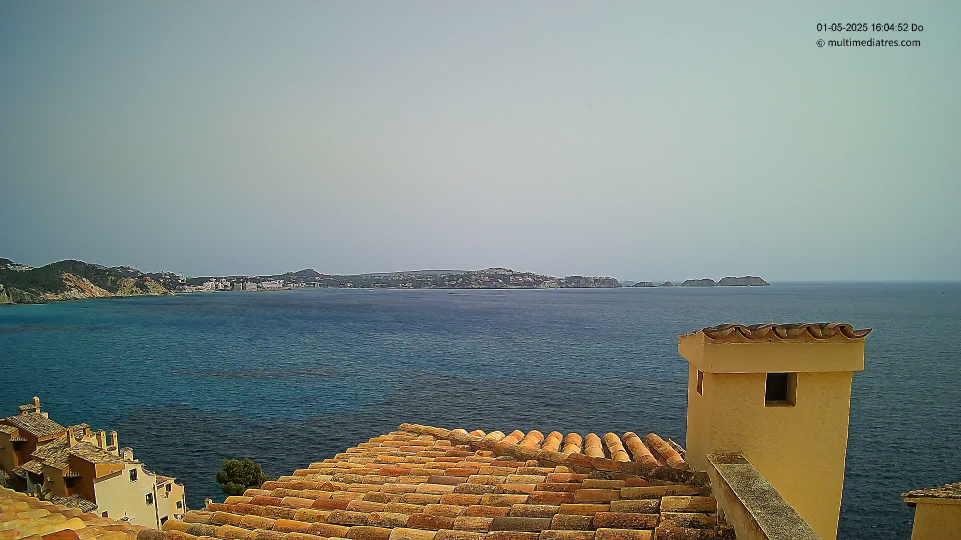 A terracotta roof overlooks a calm, blue sea and distant coastline under a hazy sky.