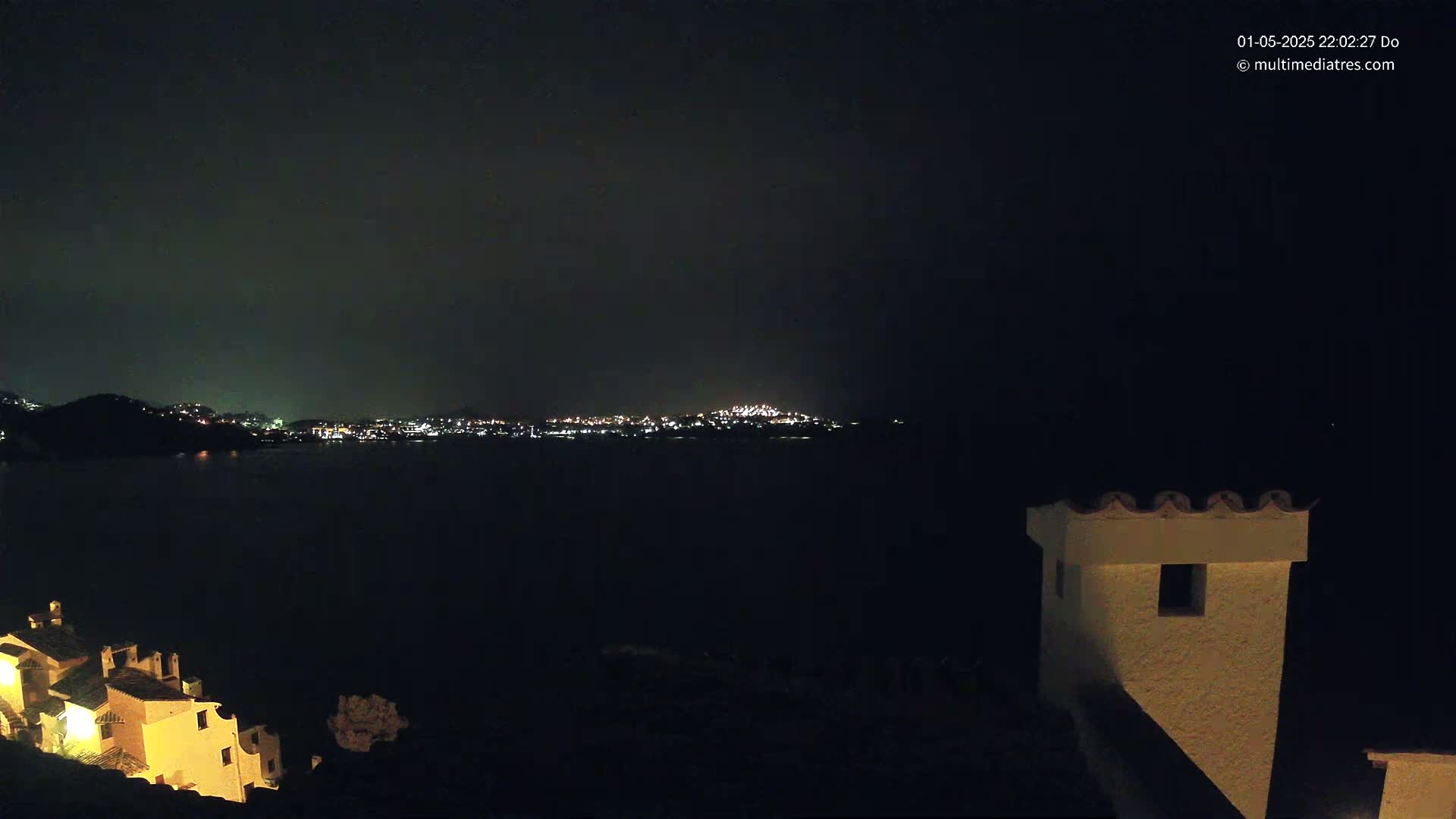 A nighttime view across dark water shows a distant city skyline illuminated by lights, with a portion of a building, possibly a chimney, in the foreground.