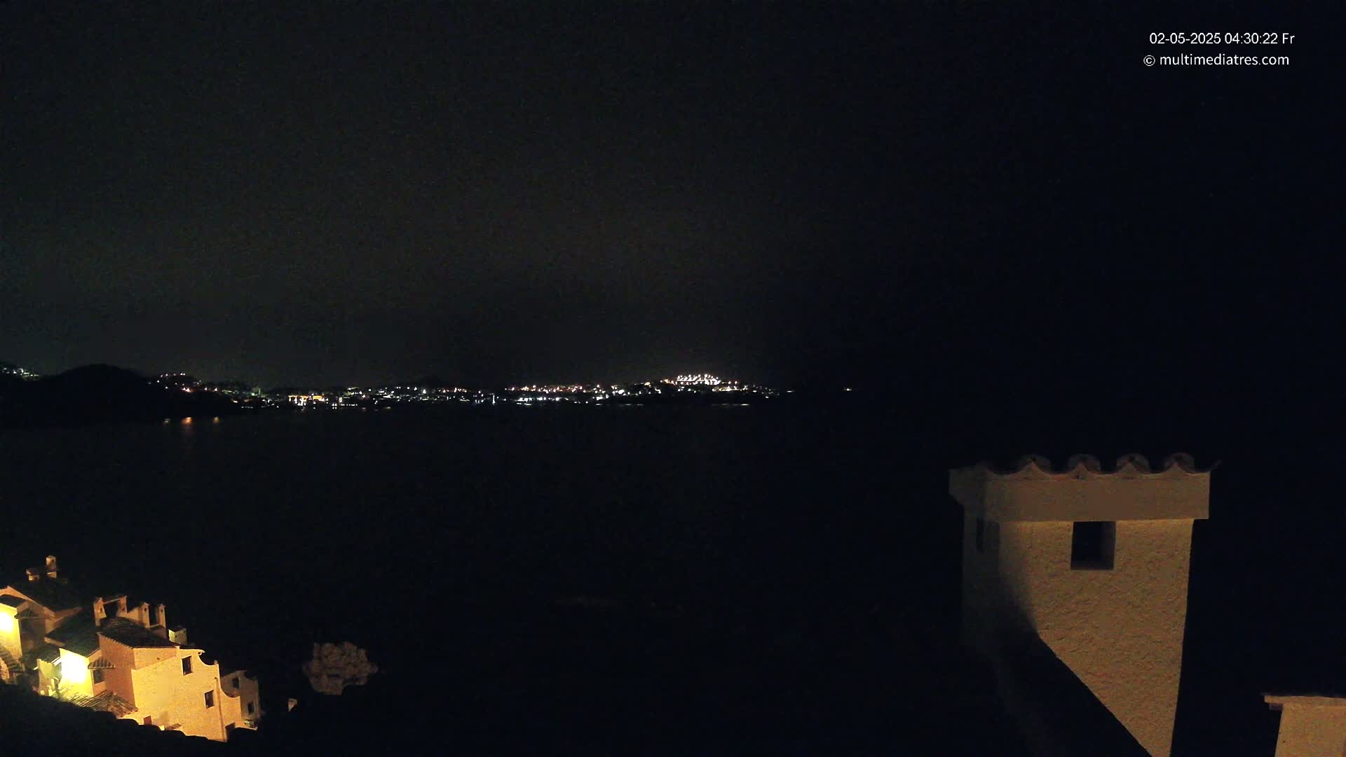 A nighttime view of a city across a dark body of water, with a building and its rooftop visible in the foreground under a clear, dark sky.