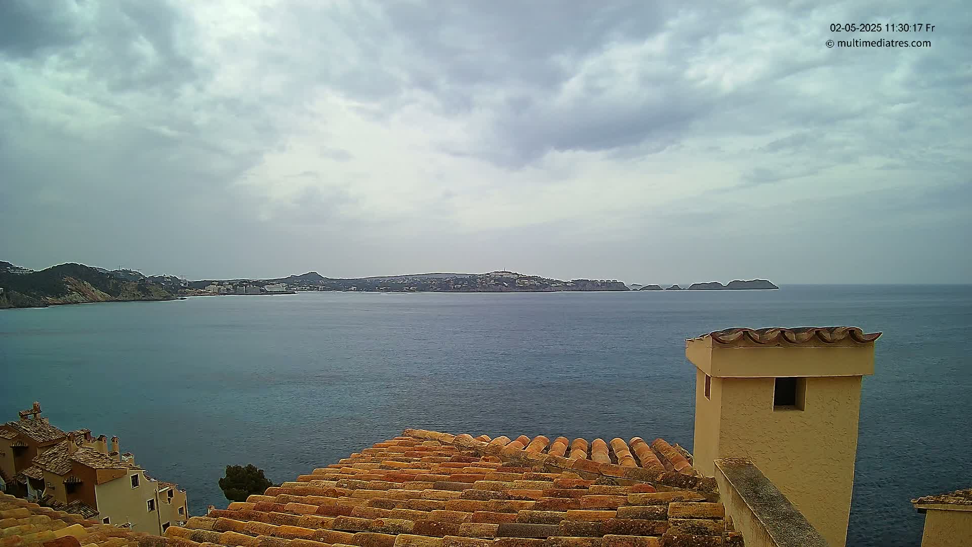 A terracotta roof overlooks a calm sea with a distant coastline under a cloudy sky.