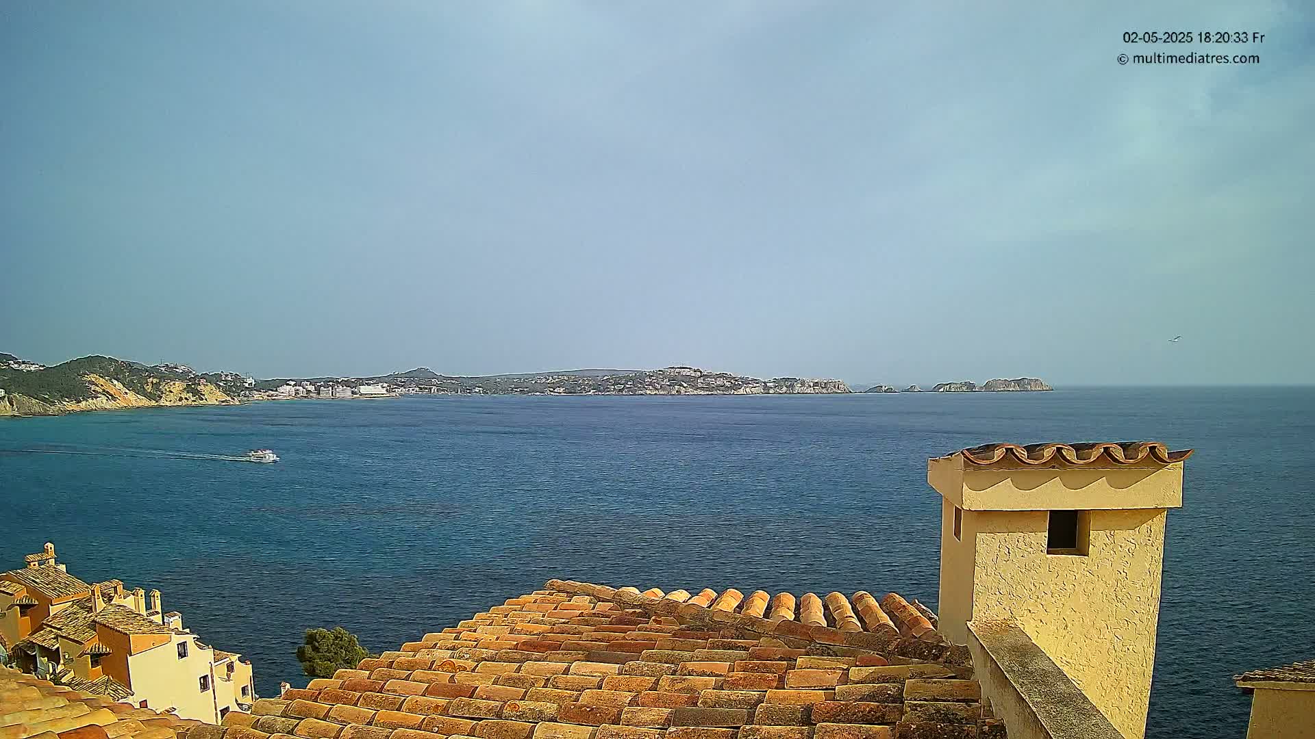 A clear, sunny day reveals a coastal scene of a calm blue sea, with distant landmasses and a closer view of terracotta rooftops and a chimney.