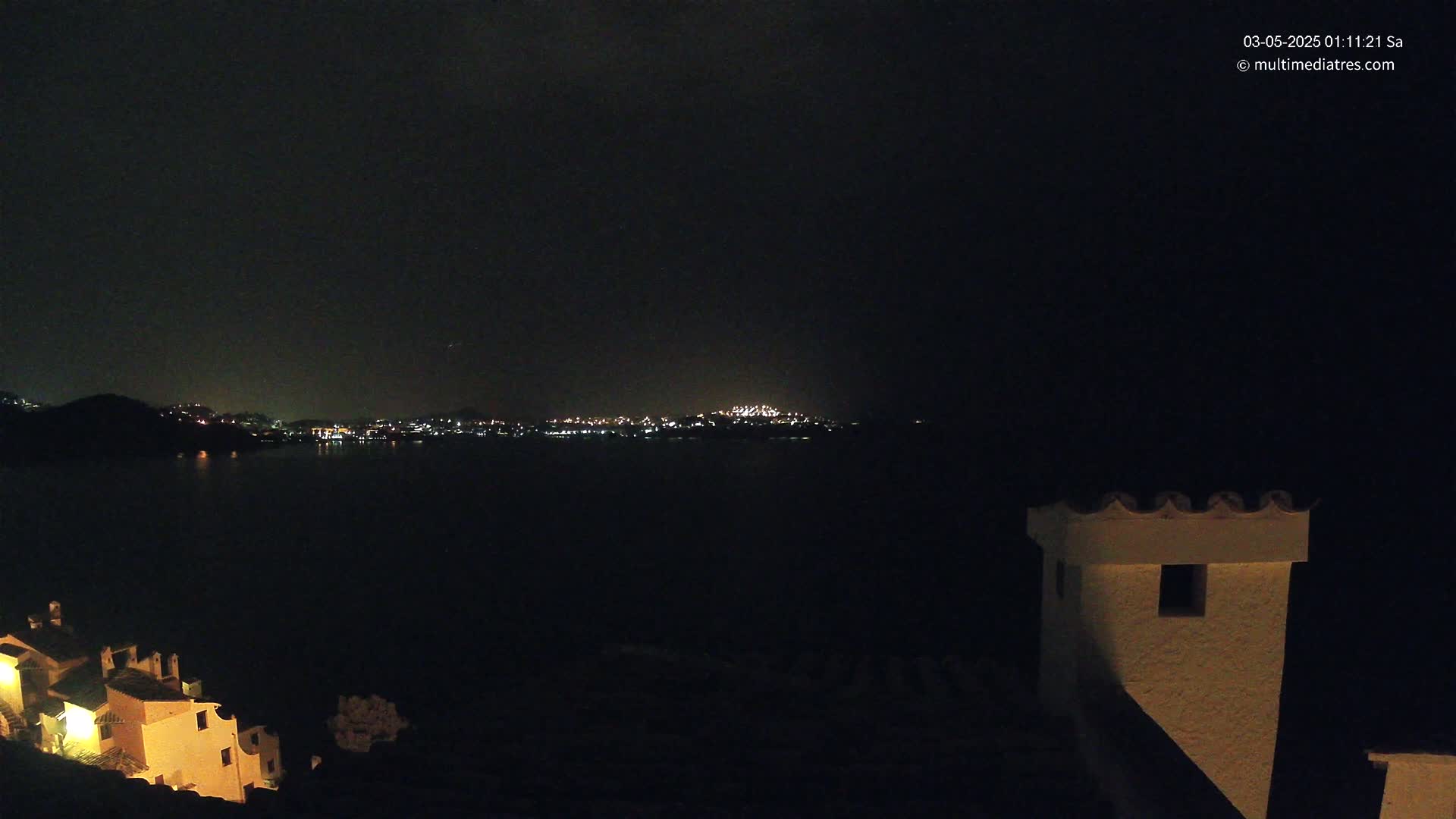 A nighttime view across a dark body of water shows a distant city skyline illuminated with lights, and closer to the foreground, a partially lit building.