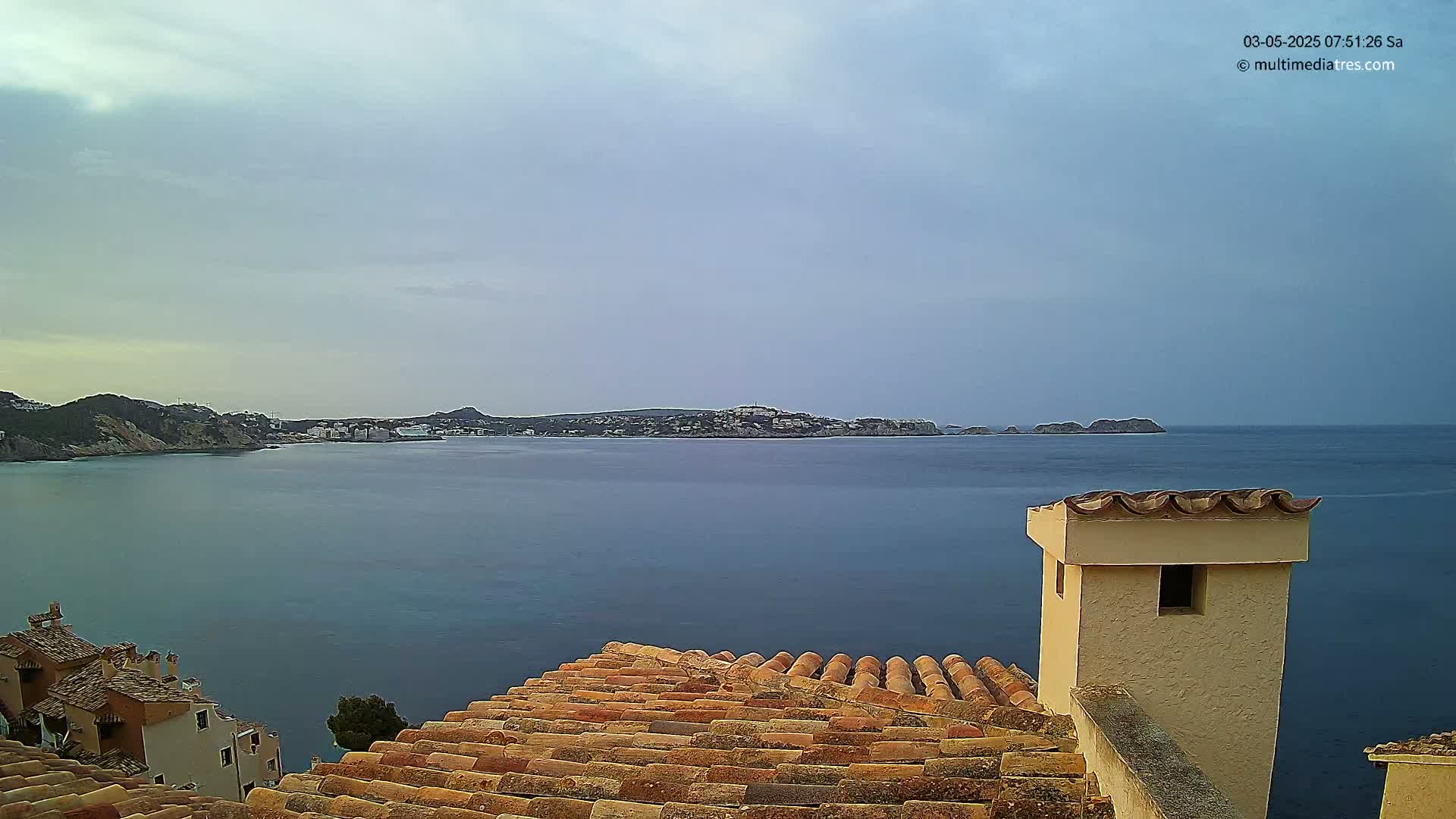 A calm, overcast day reveals a coastal view of a town nestled between a sea and distant hills, seen from a tiled rooftop with a chimney.