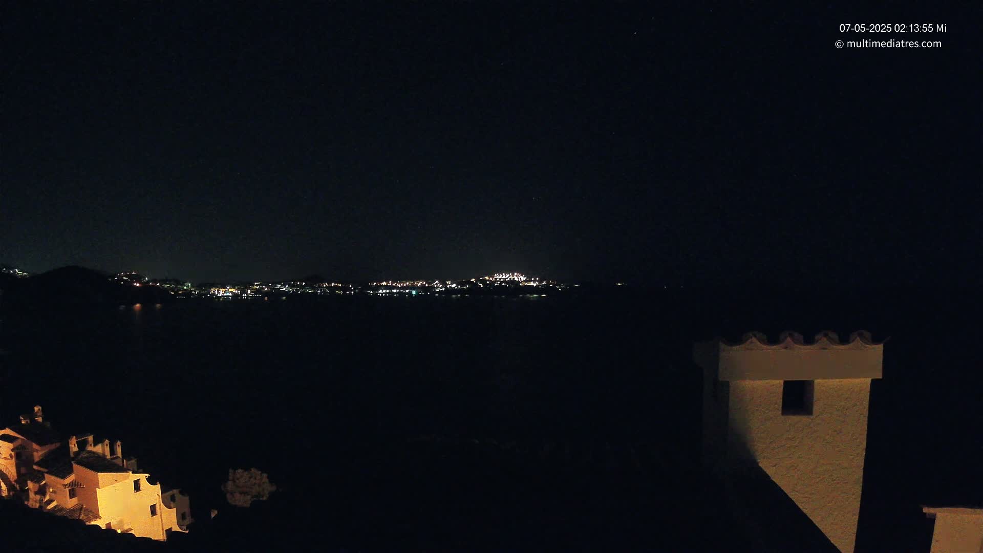 A nighttime, dark view of a city's lights across a body of water, partially obscured by darkness, with a building in the foreground.
