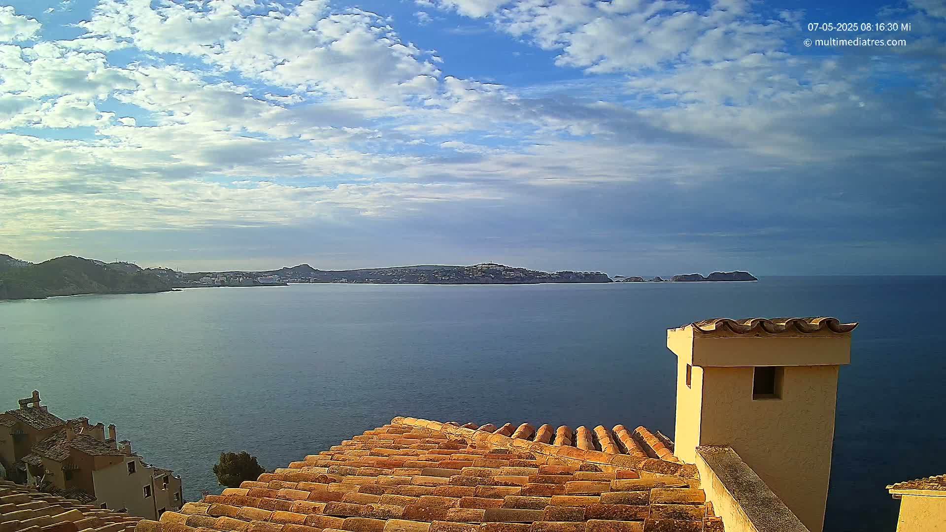 A partly cloudy sky overlooks a calm sea with a distant town and coastline visible from a terracotta roof with a beige chimney.