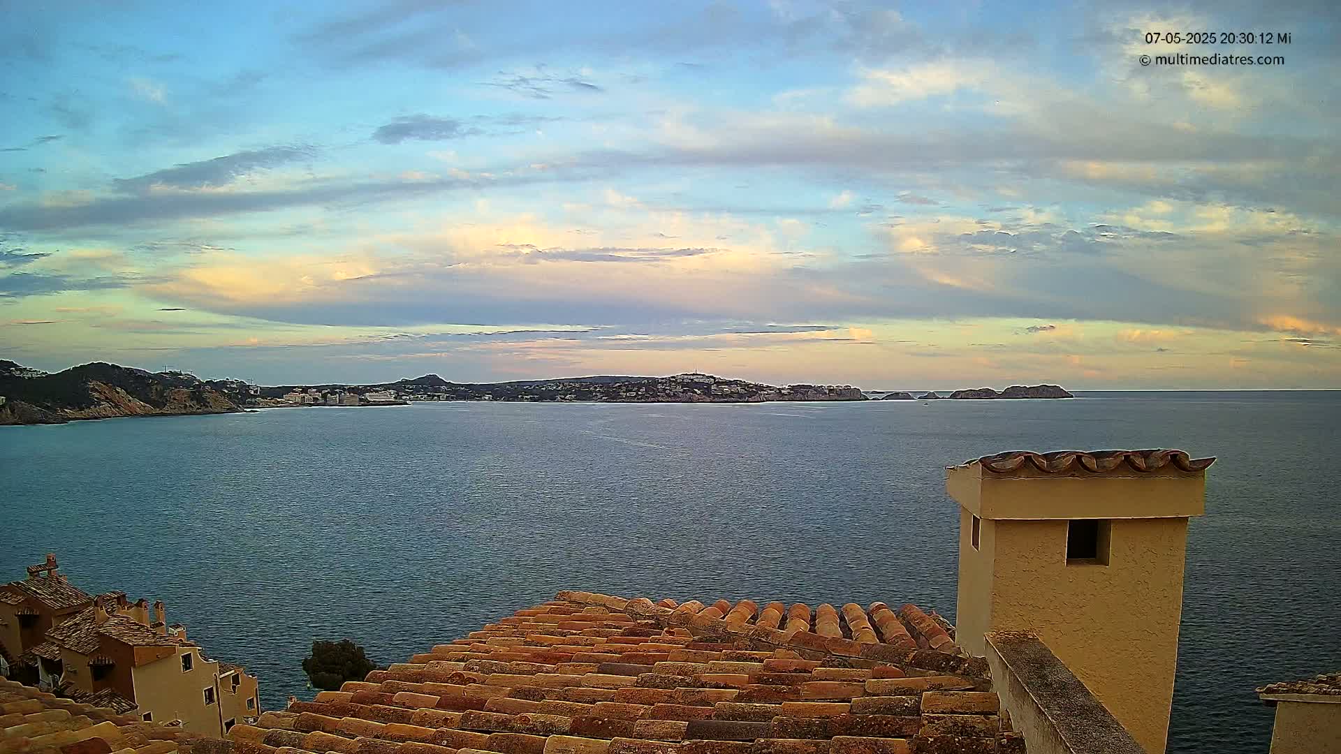 A calm sea with a distant town and islands under a partly cloudy sky is seen from a tiled rooftop with a chimney.