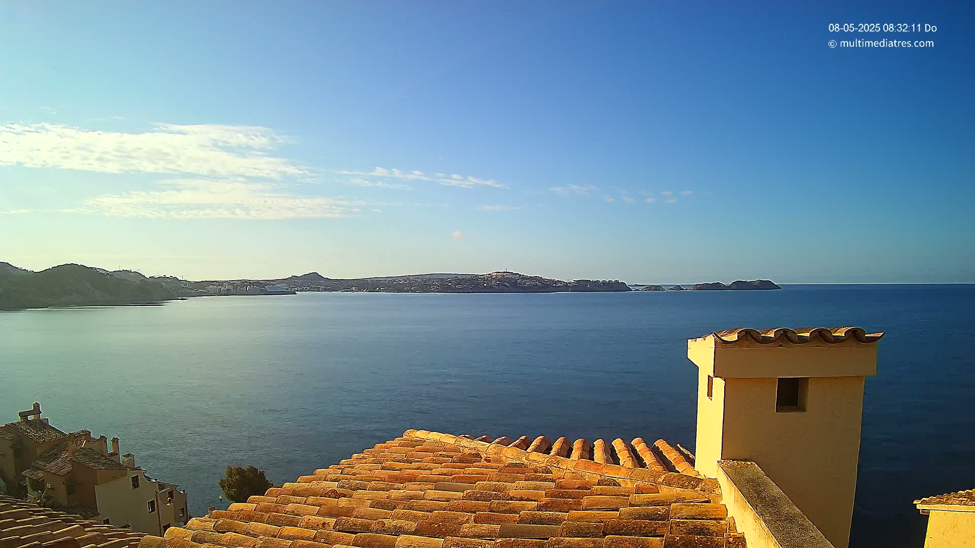 A clear, sunny day reveals a calm ocean bay, viewed from a terracotta rooftop with a tan chimney, overlooking a distant coastline.