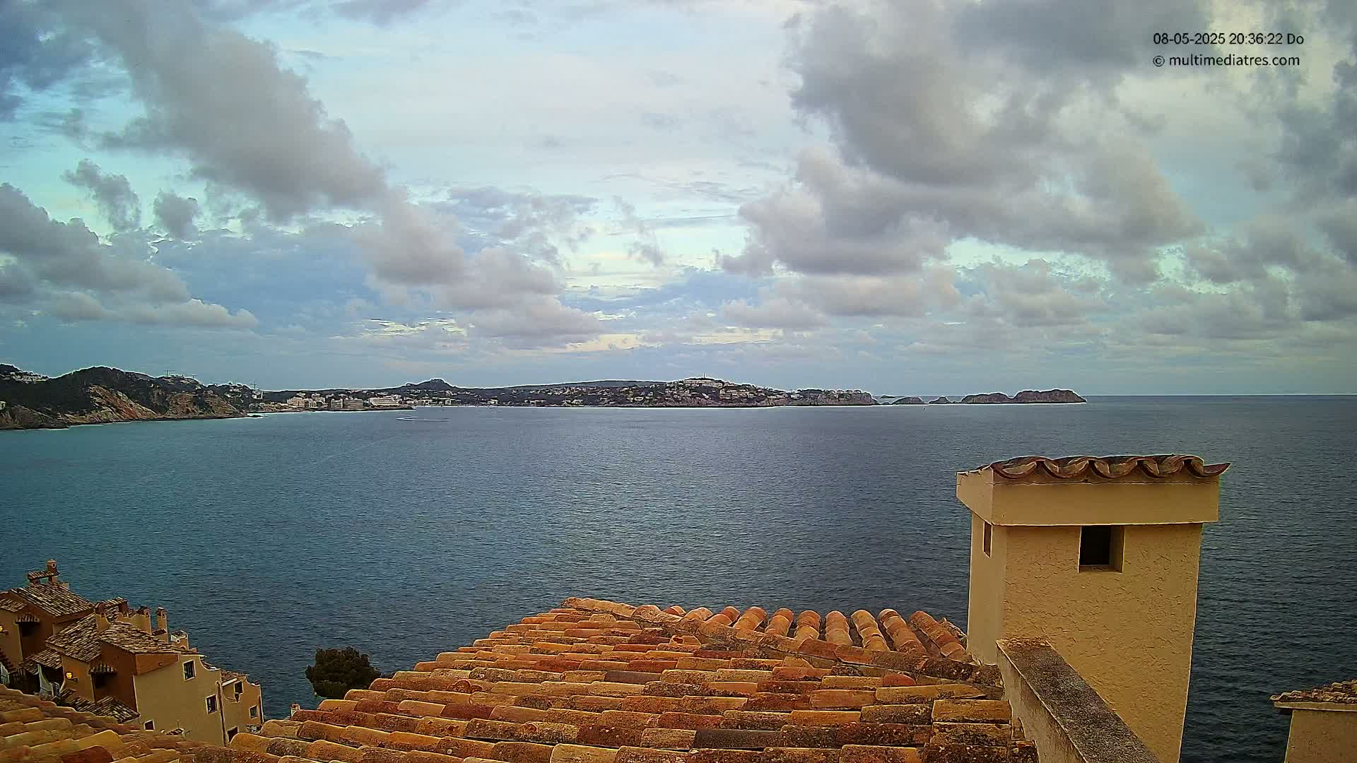 A partly cloudy sky overlooks a calm sea with a distant coastline, viewed from a tiled rooftop with a stucco chimney.