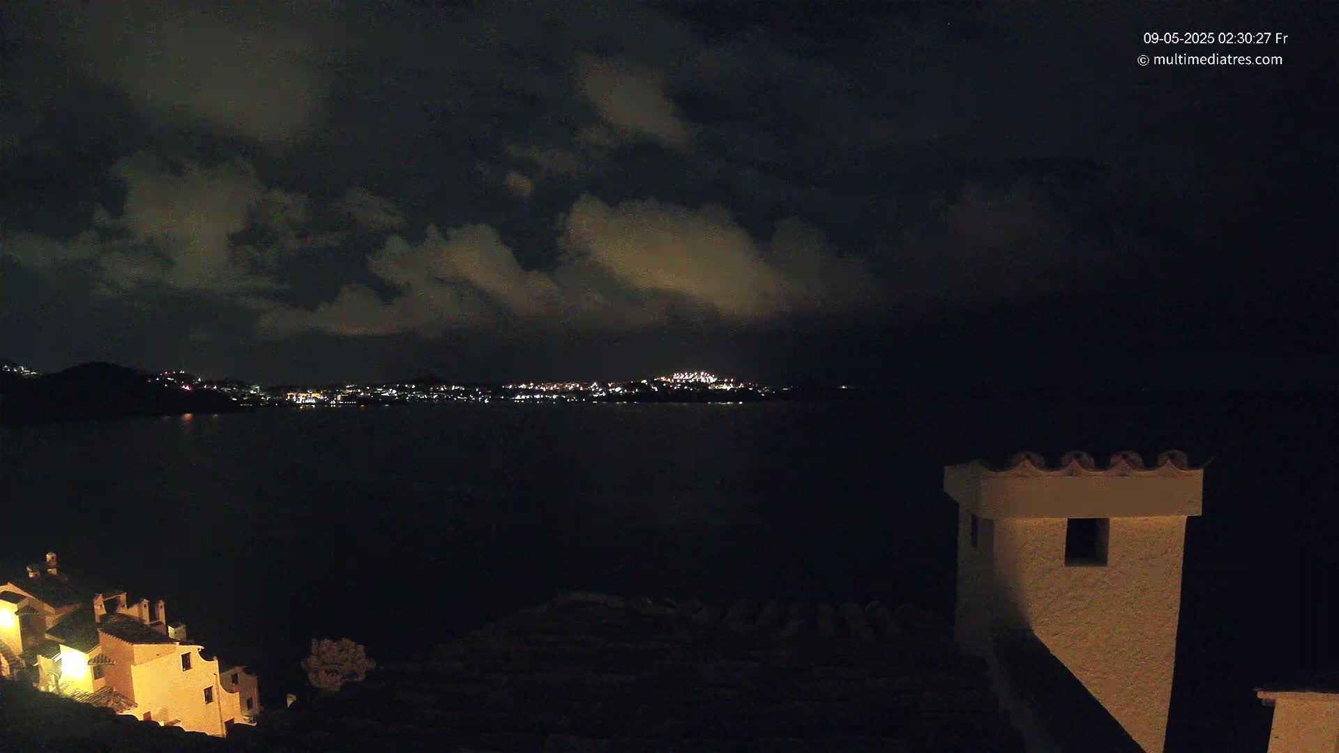 A nighttime view of a town's lights across a dark body of water, under a cloudy sky, with a portion of a building in the foreground.