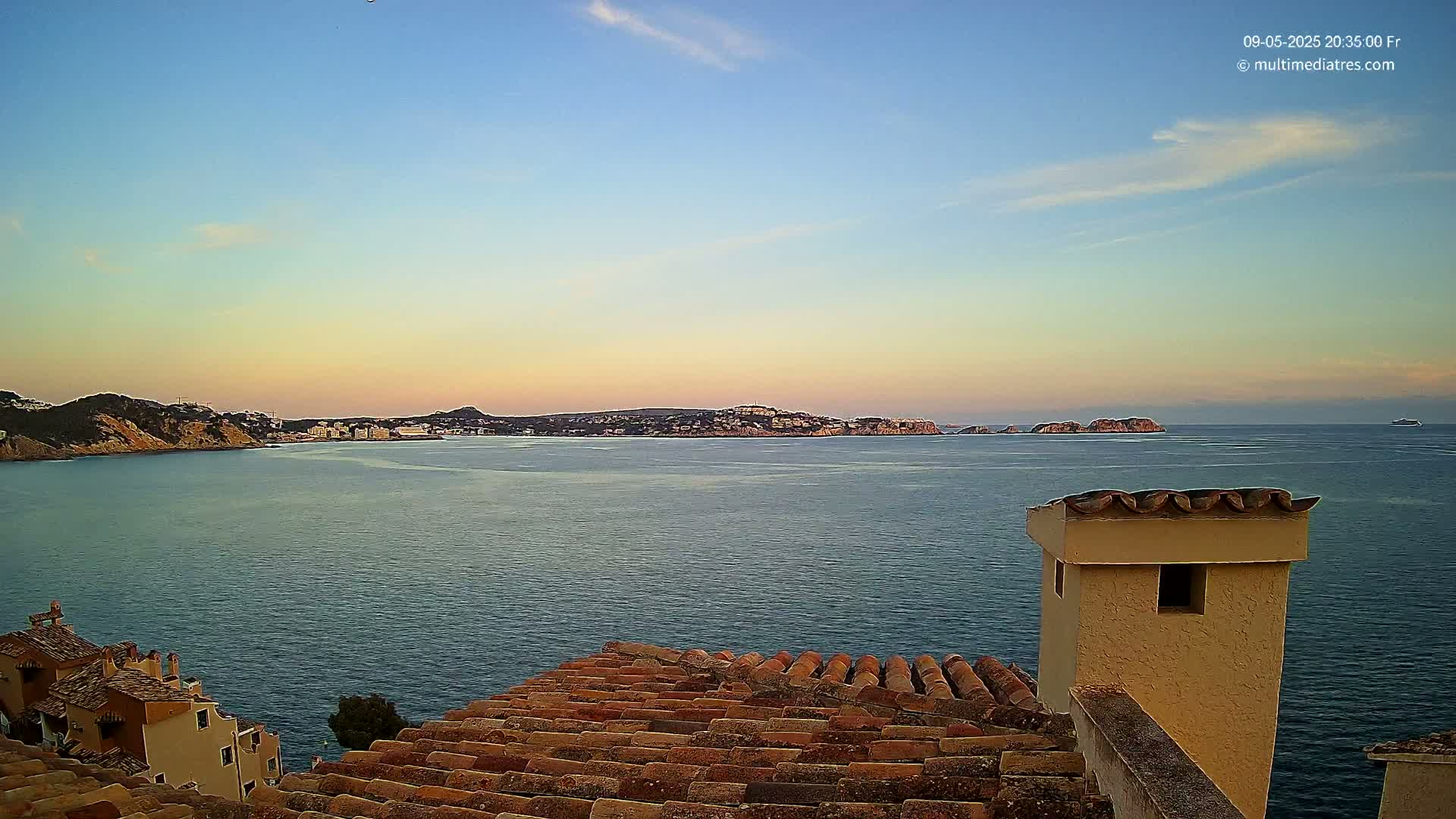 A calm sea at sunset is viewed from a tiled rooftop overlooking a coastline with buildings and distant islands.
