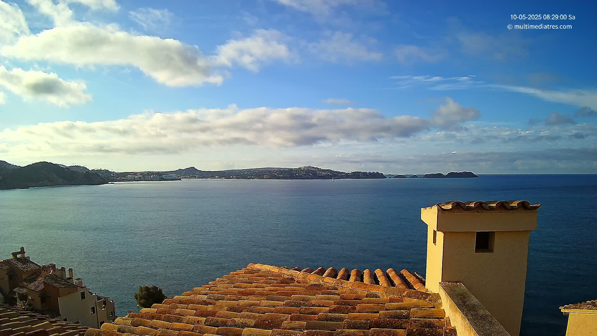 A partly cloudy sky overlooks a calm blue sea and distant coastline, viewed from a terracotta roof with a small beige chimney.