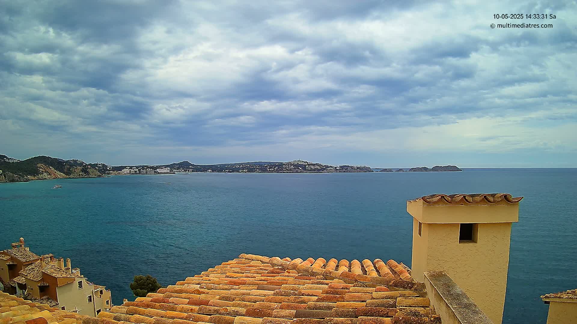 A partly cloudy sky overlooks a calm blue sea with a distant coastline and buildings, viewed from a tiled rooftop with a chimney.