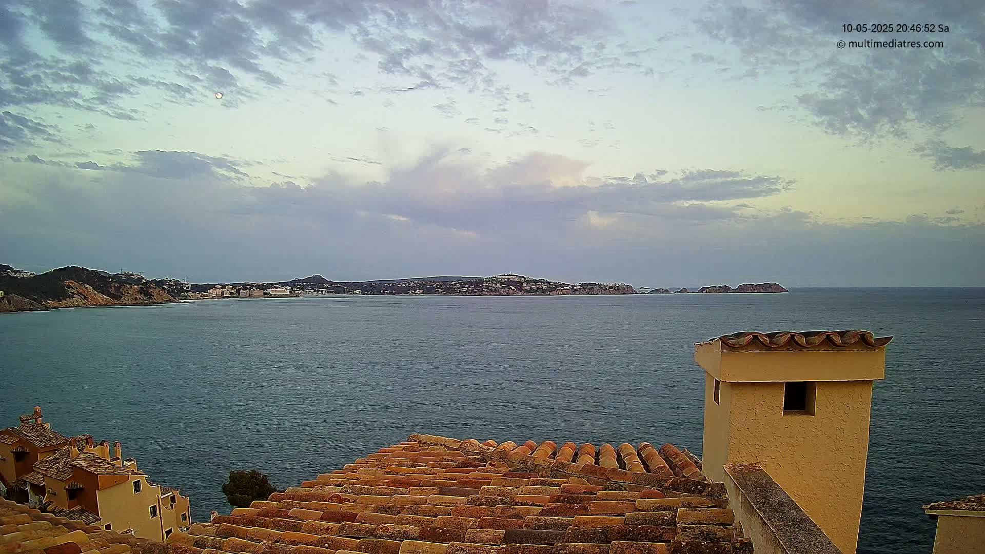 A partly cloudy sky overlooks a calm sea, with a coastal town and hills visible in the distance, seen from a rooftop with terracotta tiles and a tan chimney.