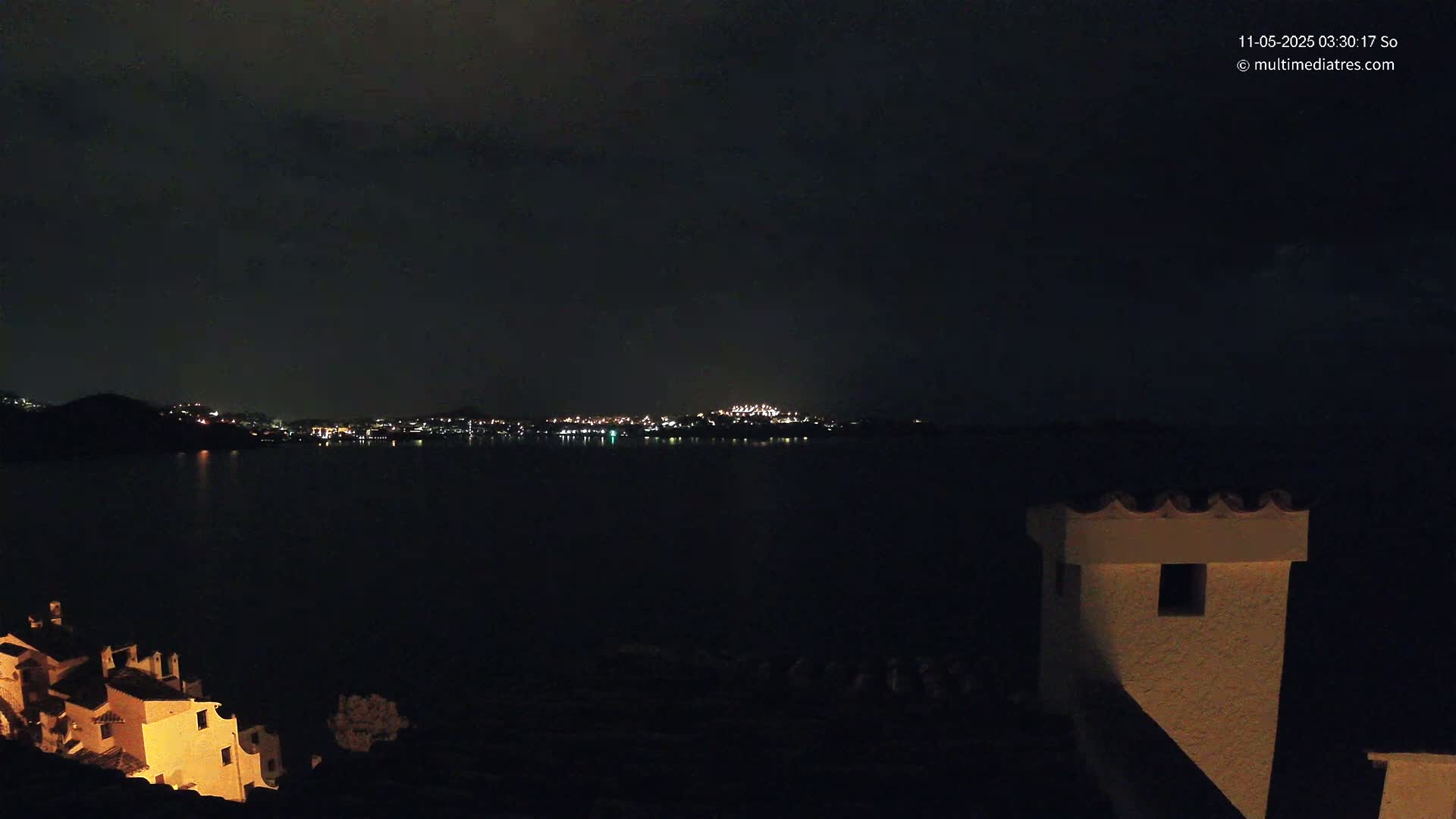 A nighttime view of a town's lights across a dark body of water, with a building partially illuminated in the foreground under a cloudy sky.