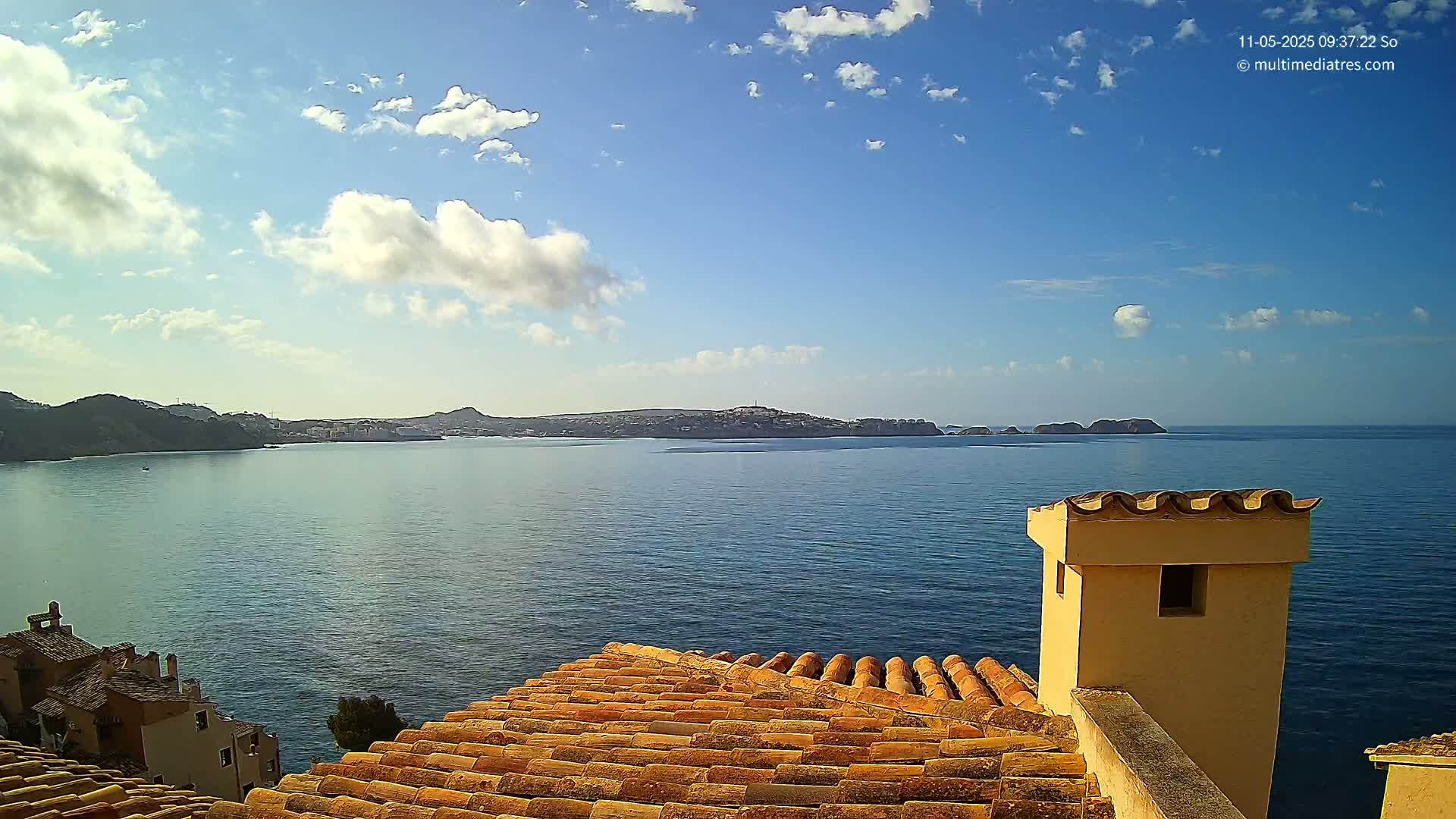 A partly cloudy blue sky overlooks a calm sea, with a distant landmass visible beyond a terracotta roof and chimney.