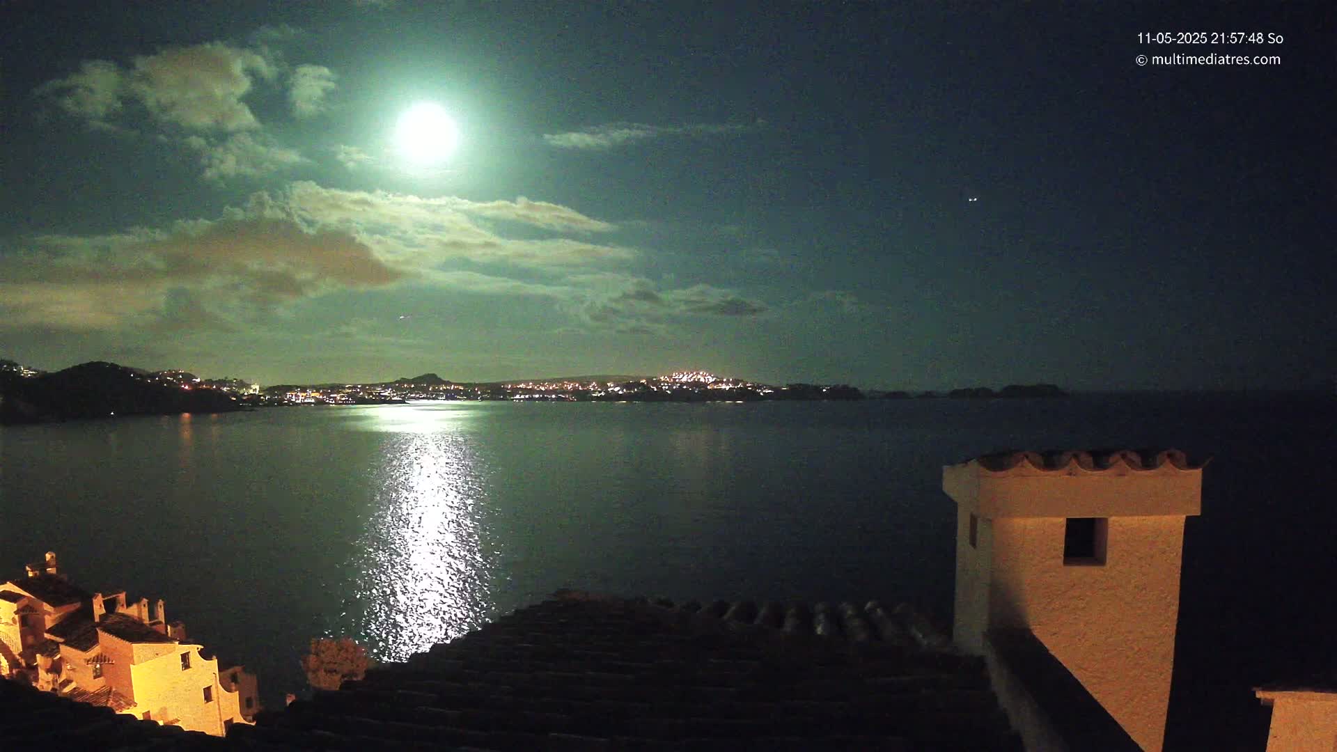 A brightly lit moon shines through partly cloudy night skies over a calm body of water reflecting the light, with a distant city skyline and buildings visible from a high vantage point.