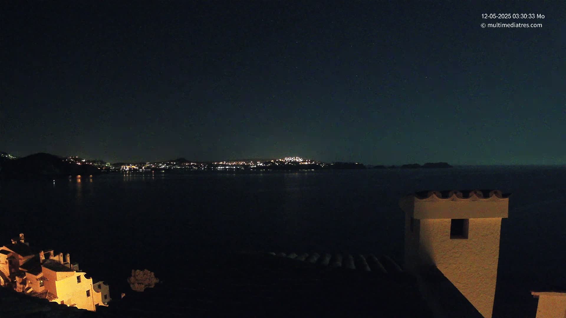 A nighttime view of a city across a dark body of water, illuminated by lights, with a building and chimney in the foreground under a clear, starry sky.