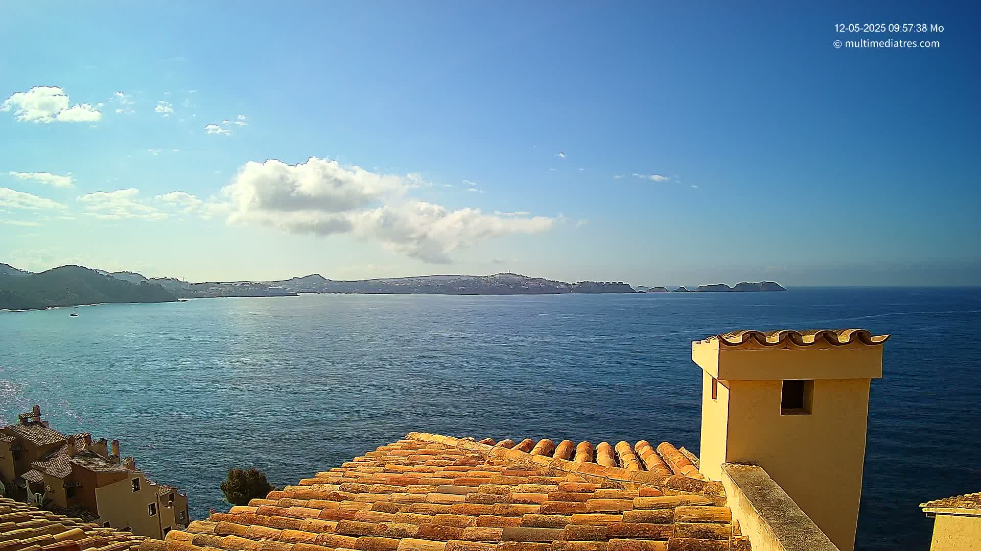 A sunny coastal view shows a calm blue sea with distant hills and islands, seen from a tiled rooftop with a light-colored chimney.