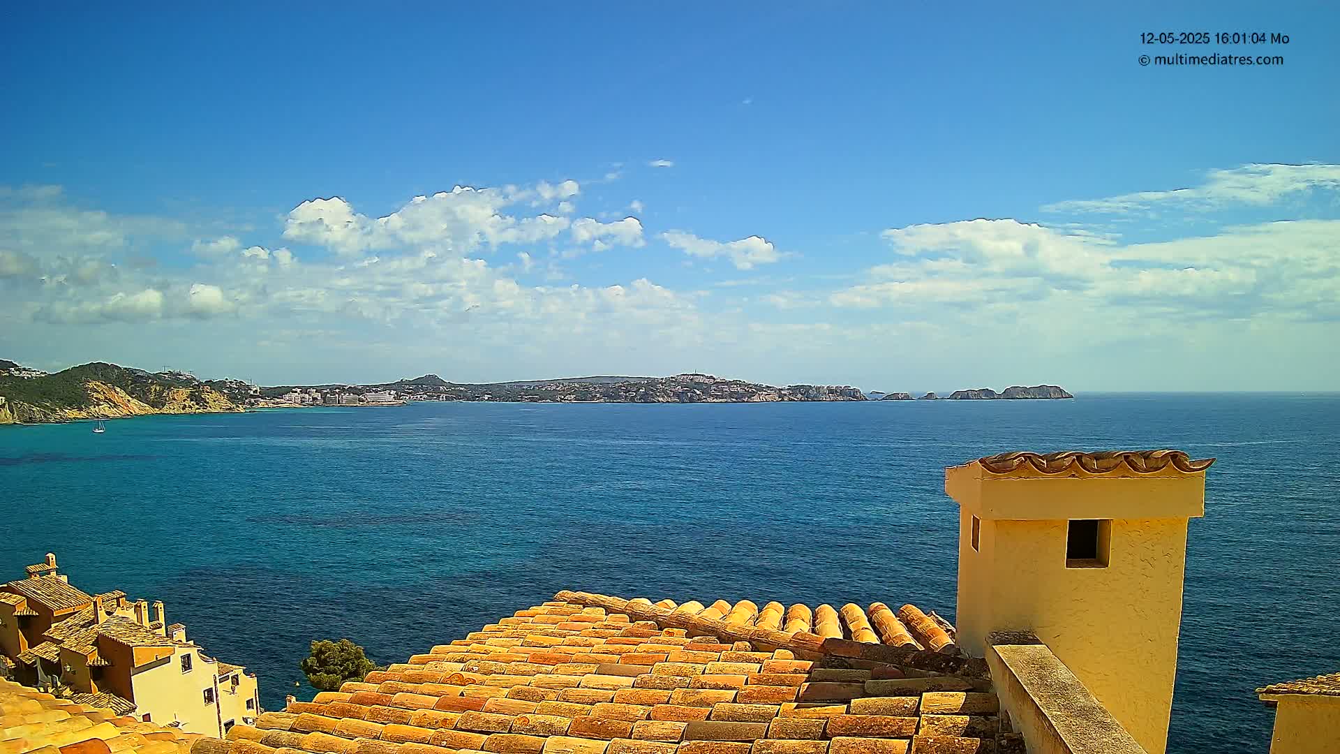 A partly cloudy blue sky overlooks a calm, blue ocean, with distant landmasses and a portion of a terracotta roof and a small, light-yellow building in the foreground.