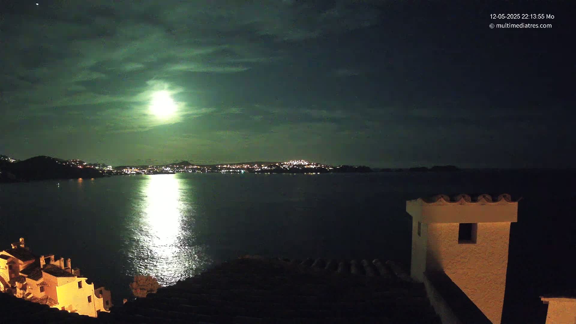 A brightly lit moon illuminates a dark, calm body of water separating a coastline of buildings from a distant city at night.