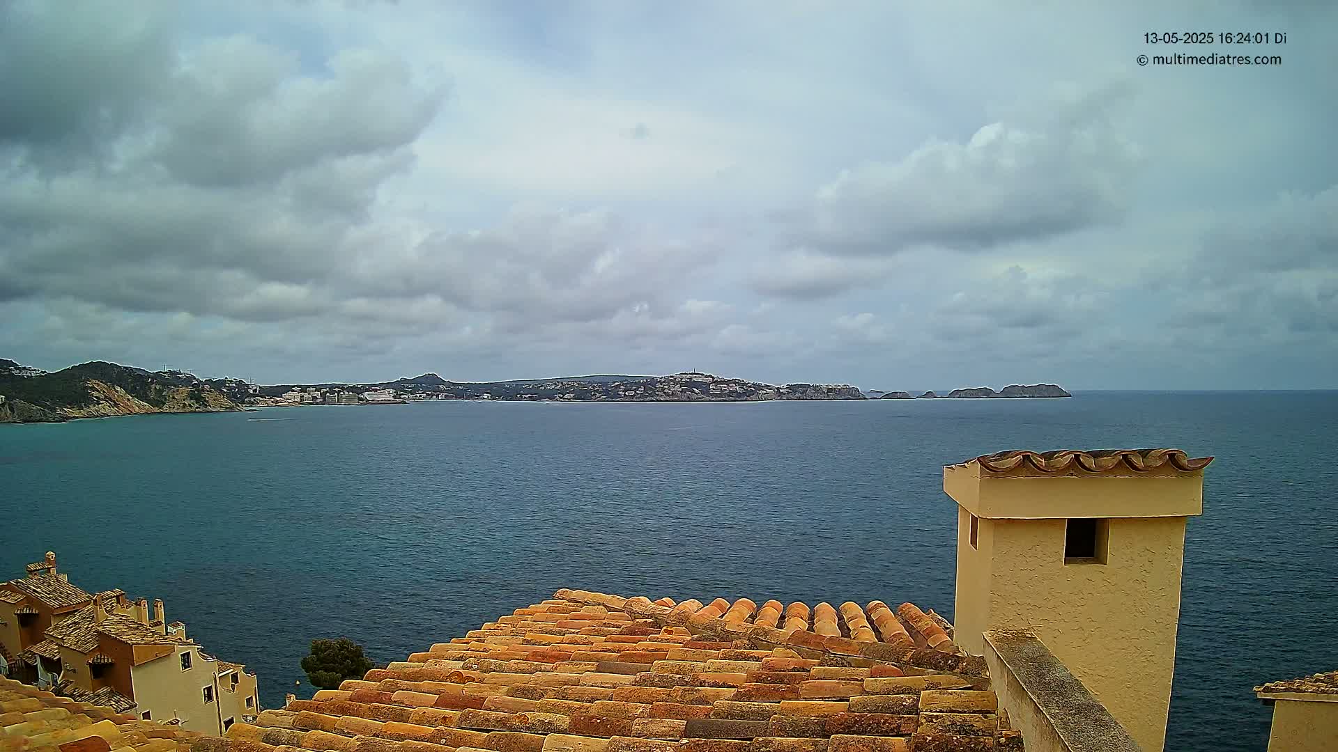 A view from a tiled rooftop shows a calm sea under a cloudy sky, with distant hills and buildings visible across the water.