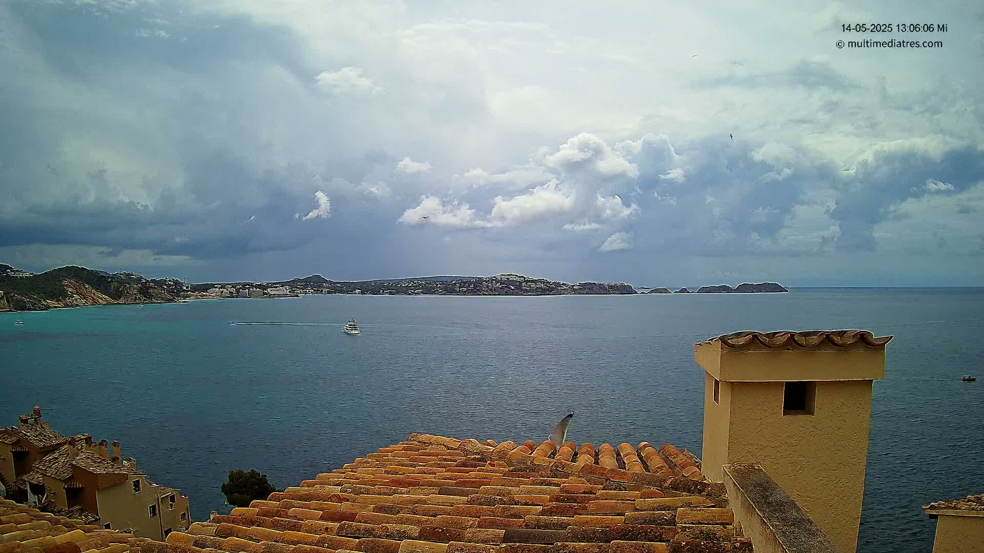 A partly cloudy sky overlooks a calm blue sea with a distant shoreline and buildings, partially visible from a terracotta roof with a tan chimney.