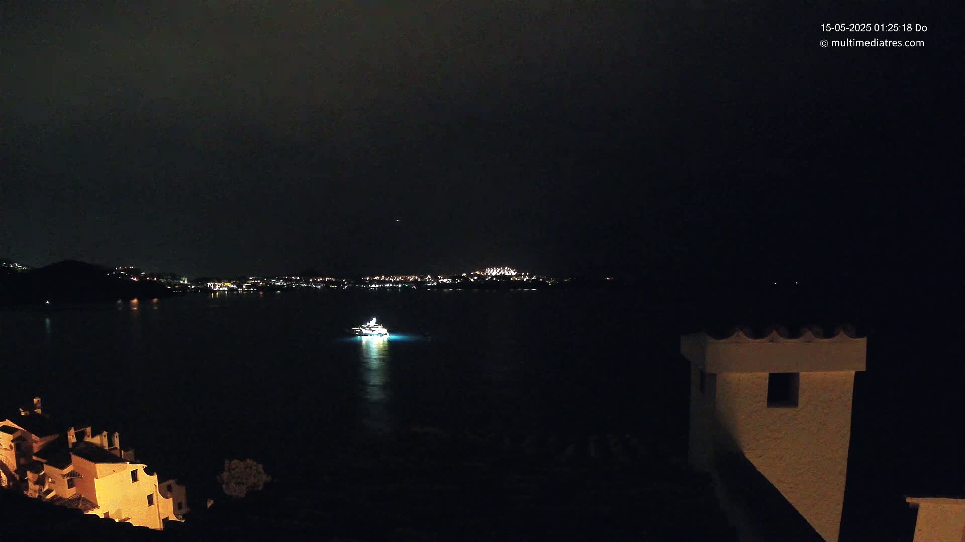 A nighttime view of a dark body of water with a single boat lit up, a distant city skyline, and a partially illuminated building in the foreground under a dark, clear sky.