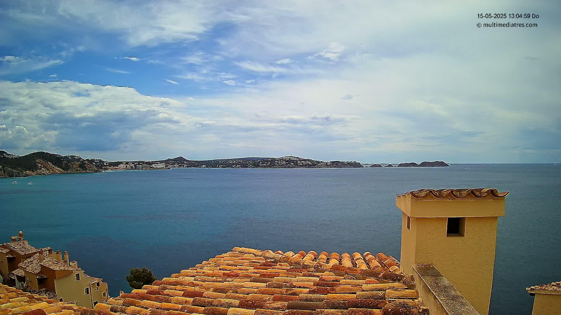 A partly cloudy sky overlooks a calm blue sea, with a distant town nestled between hills, visible from a terracotta roof and chimney.
