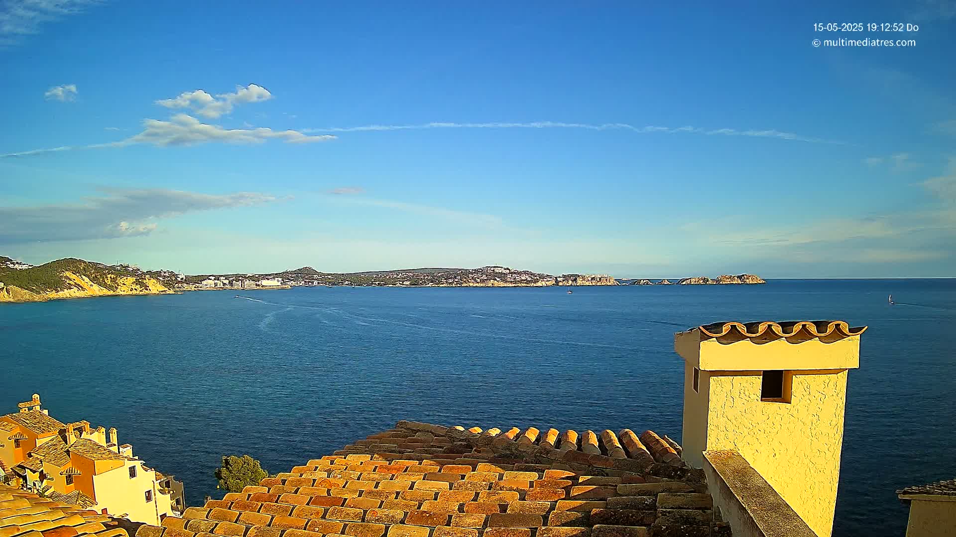 A partly cloudy blue sky overlooks a calm blue sea, with a distant coastline visible, and a view from a terracotta roof with a yellow chimney in the foreground.
