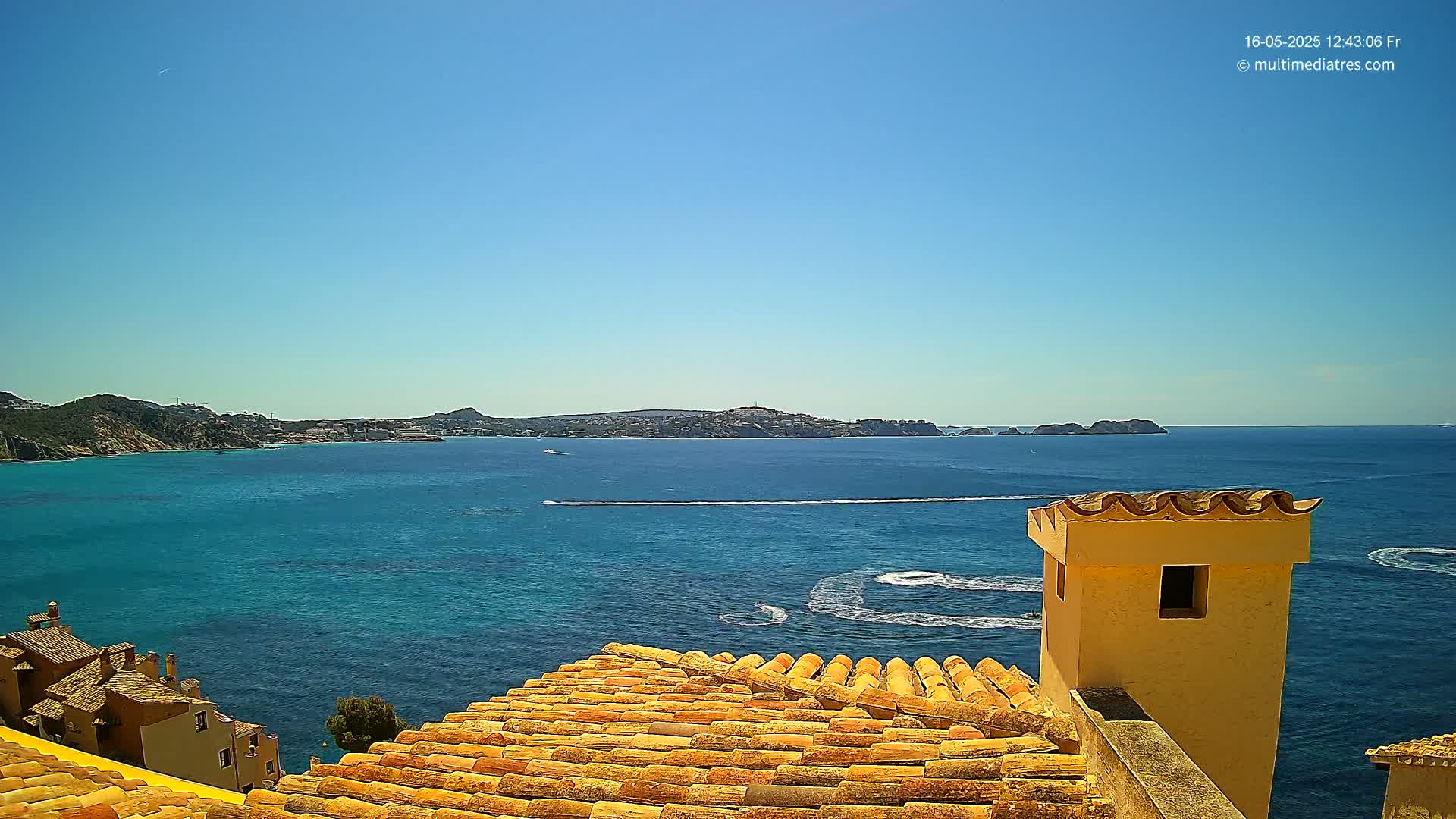 A terracotta roof overlooks a clear blue sea on a sunny day, with distant hills and small boats visible on the water.