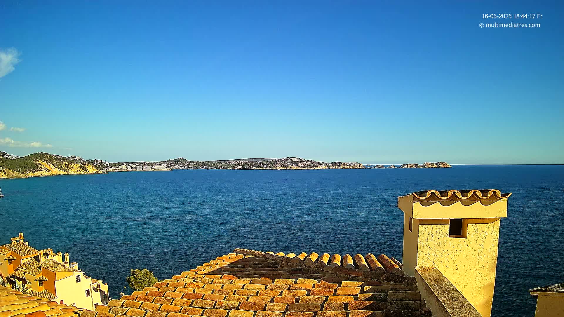 A terracotta roof overlooks a calm blue sea and distant coastline under a clear blue sky.