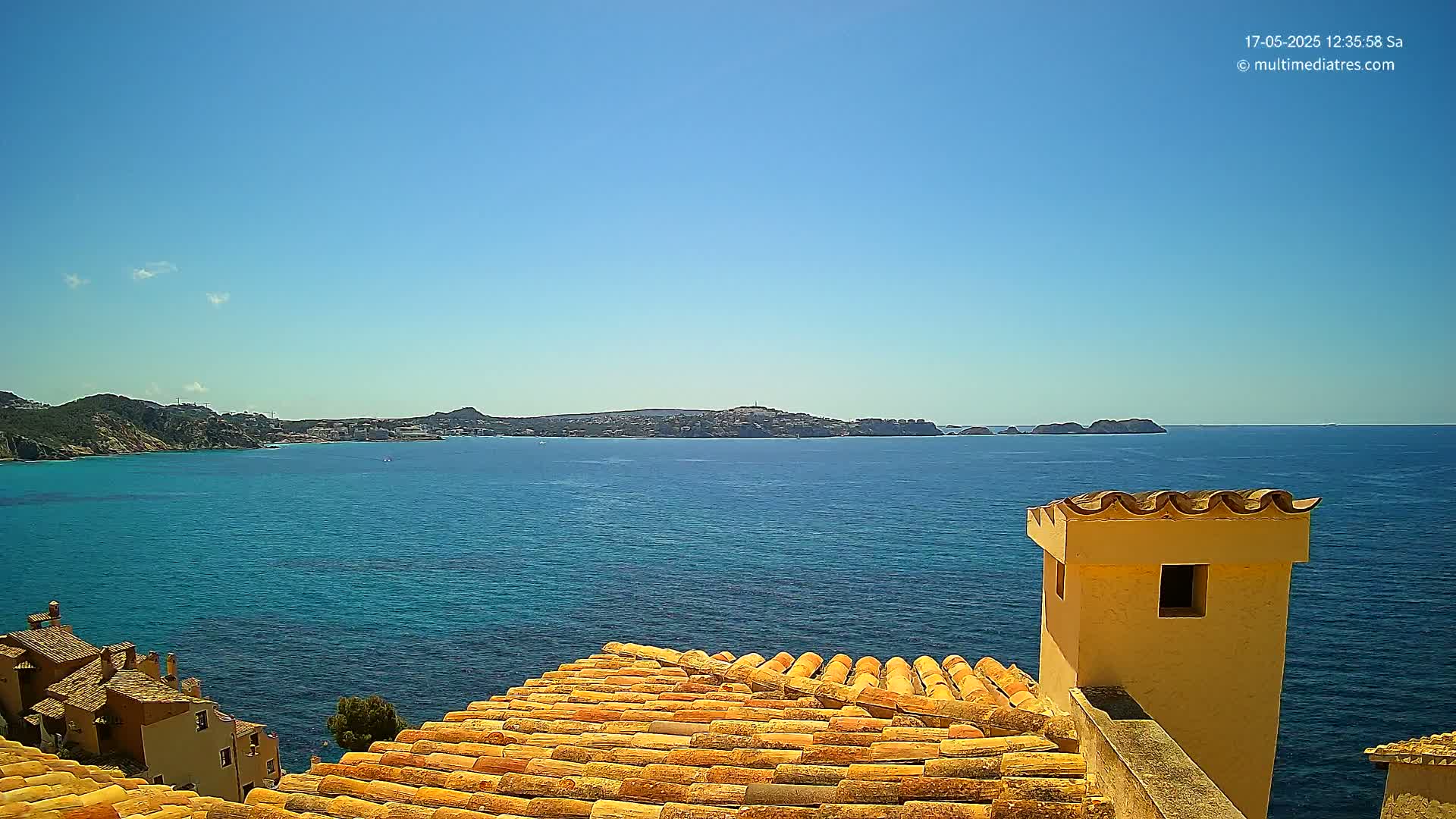 A clear blue sky overlooks a calm, blue ocean and a terracotta-tiled roof, with a distant coastline visible in the background.