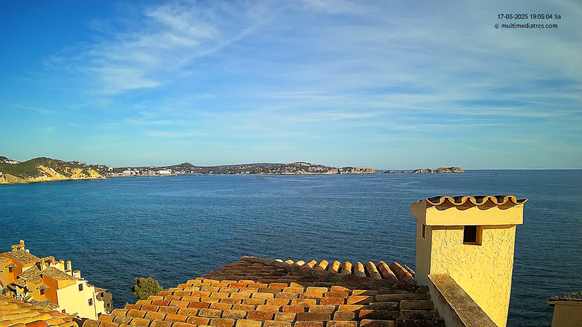 A view from a terracotta roof shows a calm blue sea, a distant coastline with buildings, and a partly sunny sky.
