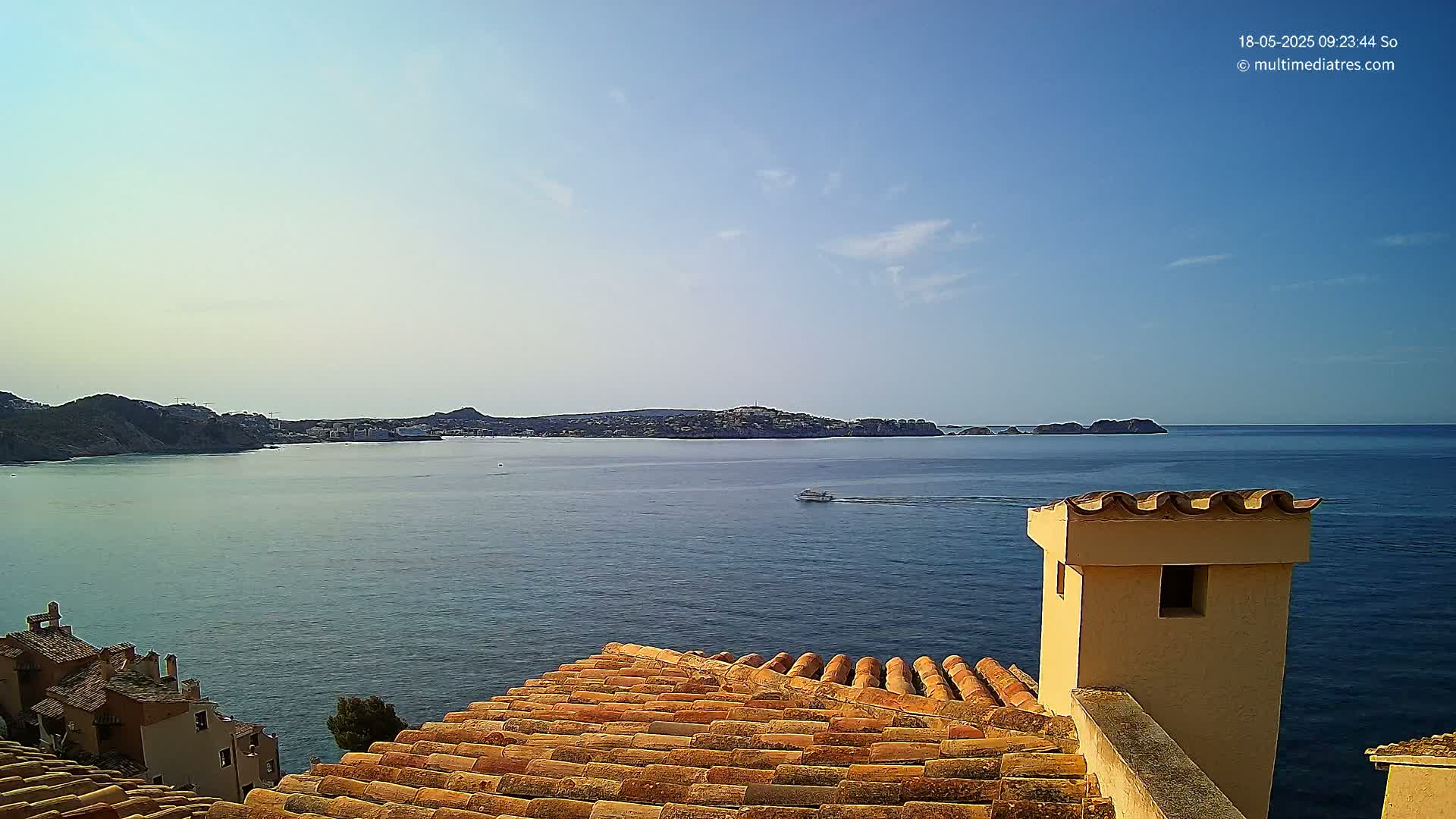 A terracotta roof overlooks a calm blue sea and distant coastline under a clear sky.