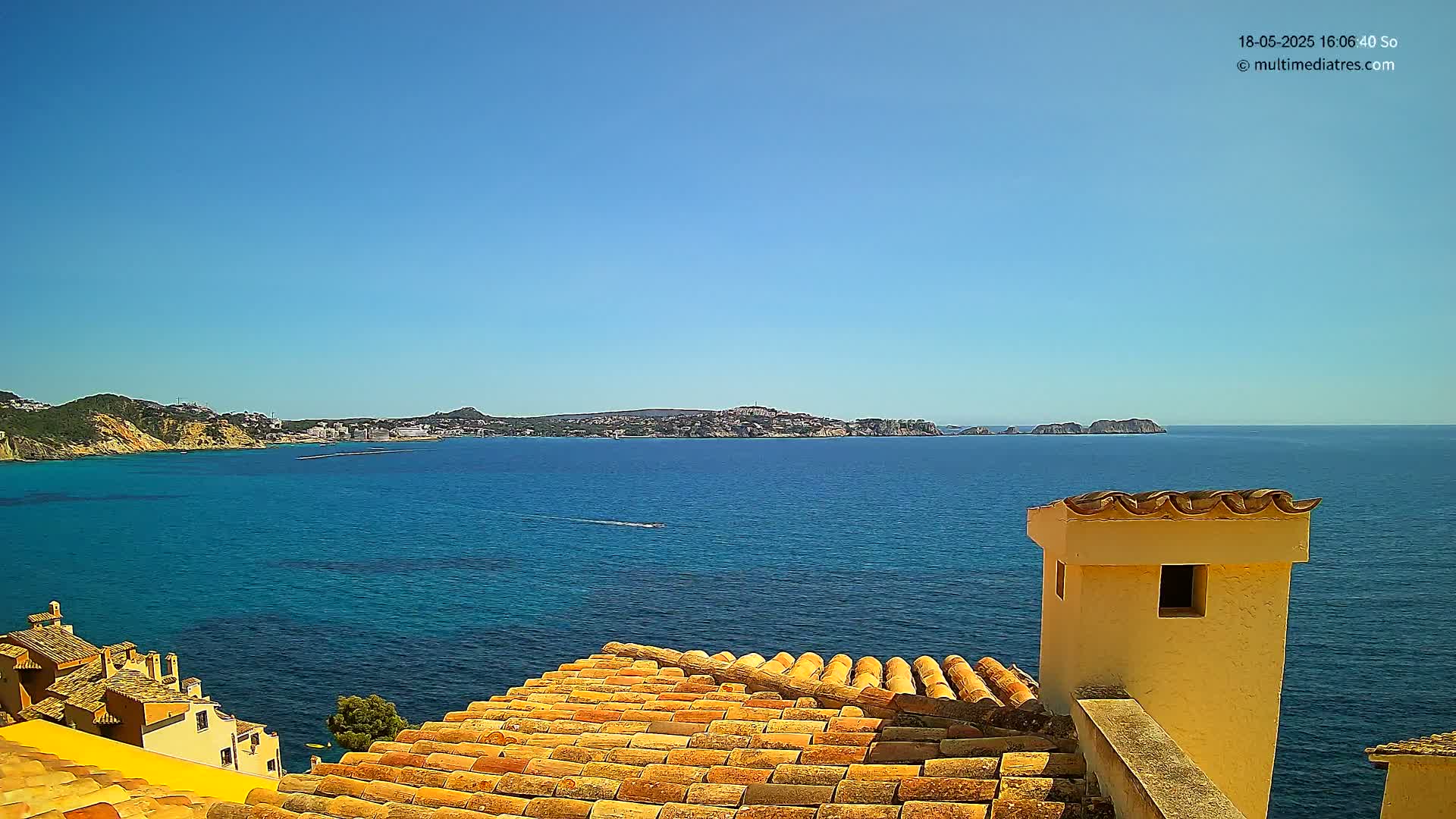 A terracotta roof overlooks a calm, blue sea and distant coastline under a clear, sunny sky.