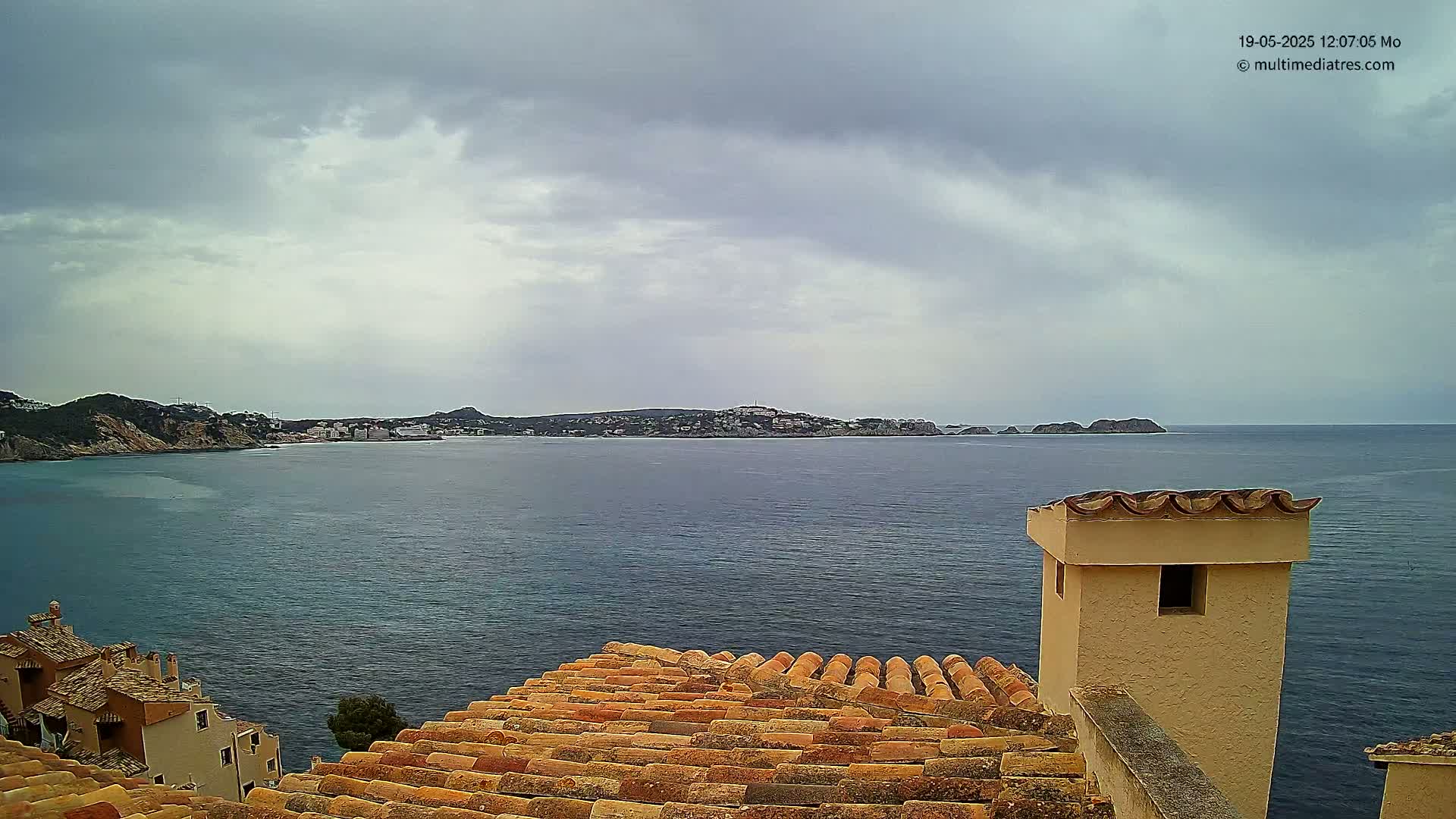 A view from a tiled rooftop shows a calm sea under a cloudy sky, with a distant coastline featuring buildings and hills.