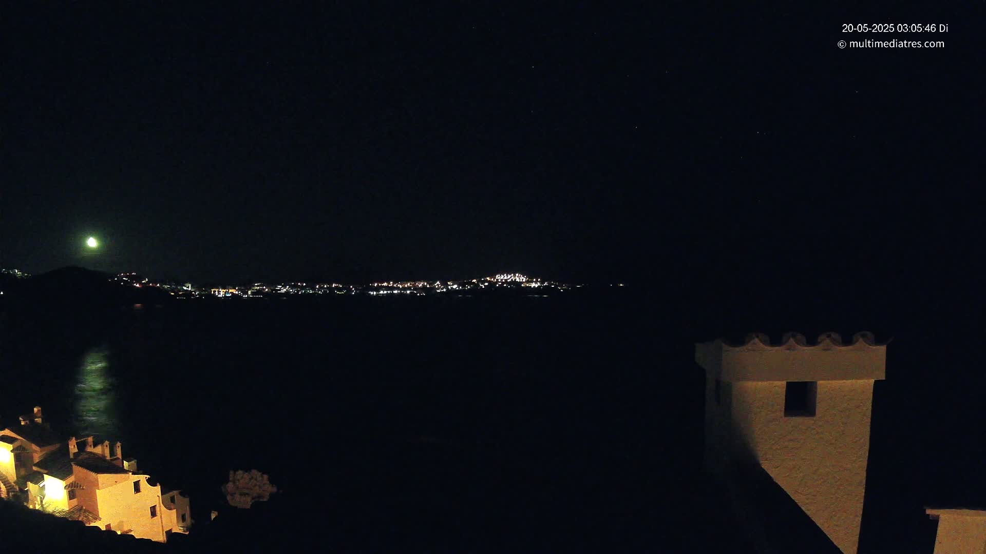 A nighttime coastal scene shows a moonlit body of water separating a collection of illuminated buildings from another larger cluster of lights across the water, with a building’s chimney in the foreground.