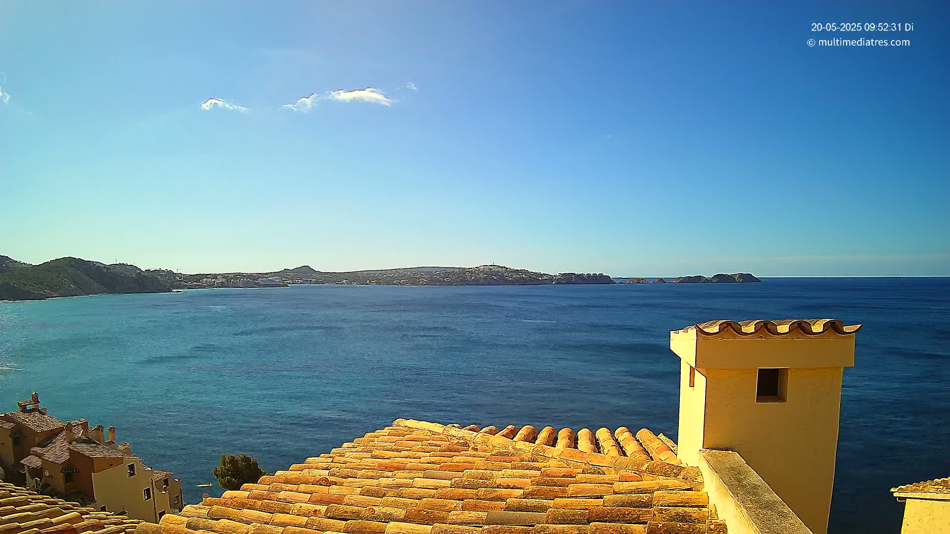 A terracotta roof overlooks a calm, blue sea and distant coastline under a mostly clear sky.