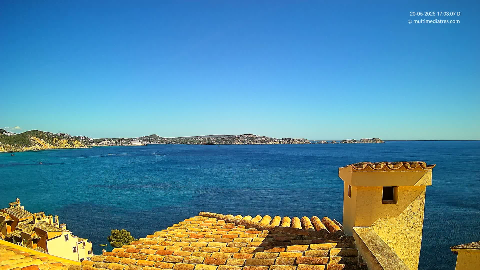 A tiled roof overlooks a clear blue sea with distant landmasses under a bright, sunny sky.